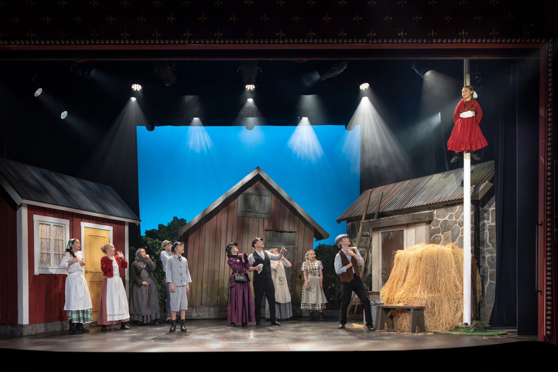 Scenbild med ensemblen från Emil i Lönneberga på Intiman i Stockholm. Ida är upphissad i flaggstången medan ensemblen står nedanför och tittar upp mot henne.