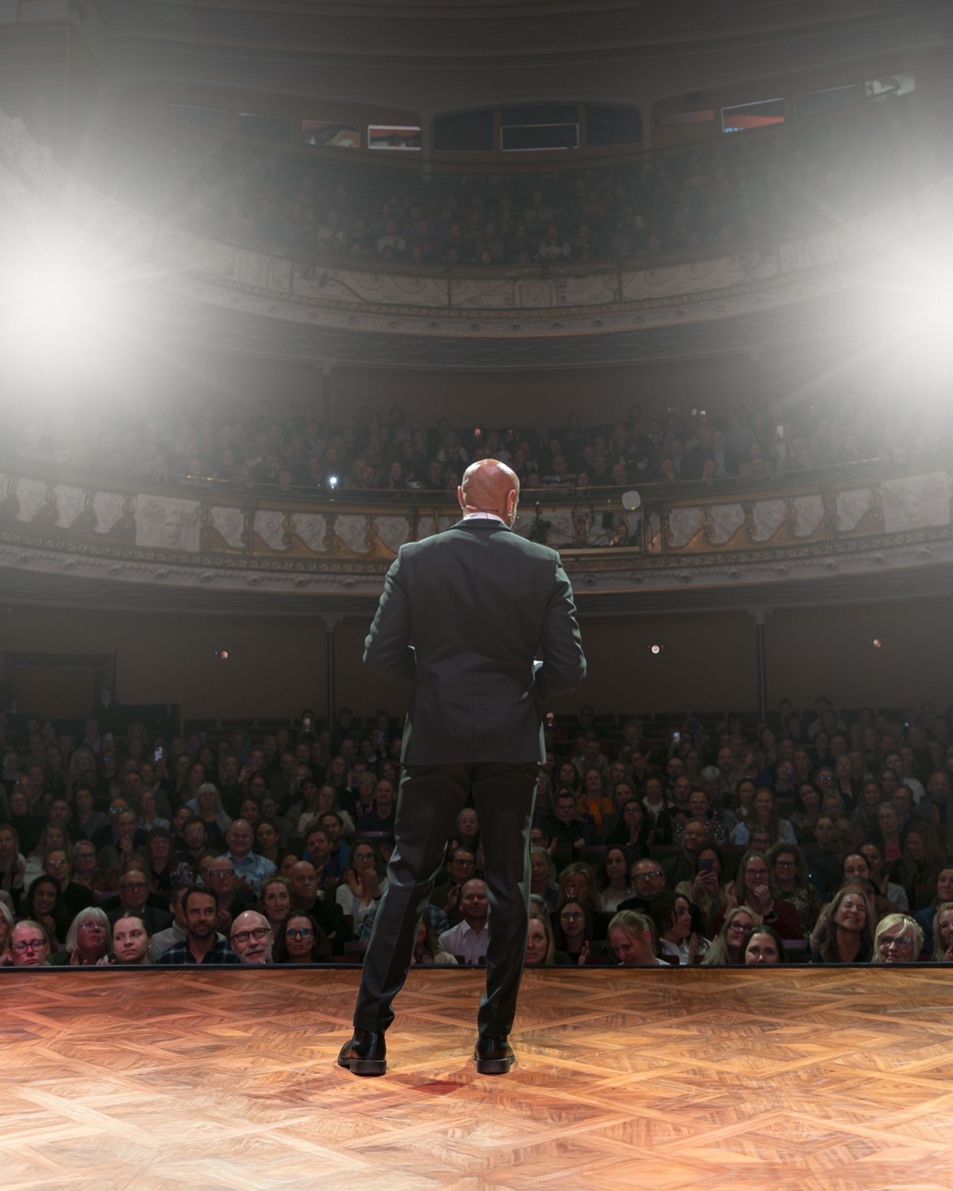 Manér för All Inclusive - Aaron Kroon med gäster. Aaron står på scen med kostym och tittar ut över en publik. Bilden är tagen bakifrån
