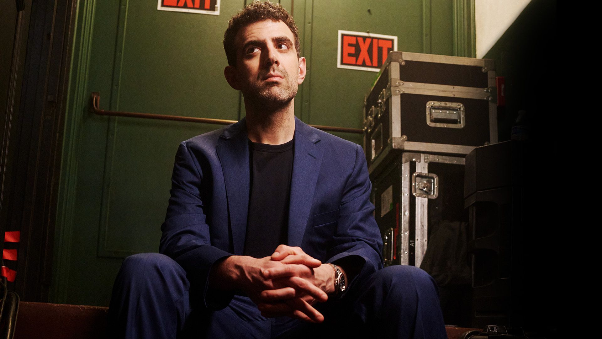 Sam Morril standup show – komiker i blå kostym bakom scen med EXIT-skyltar, porträtt inför liveföreställning