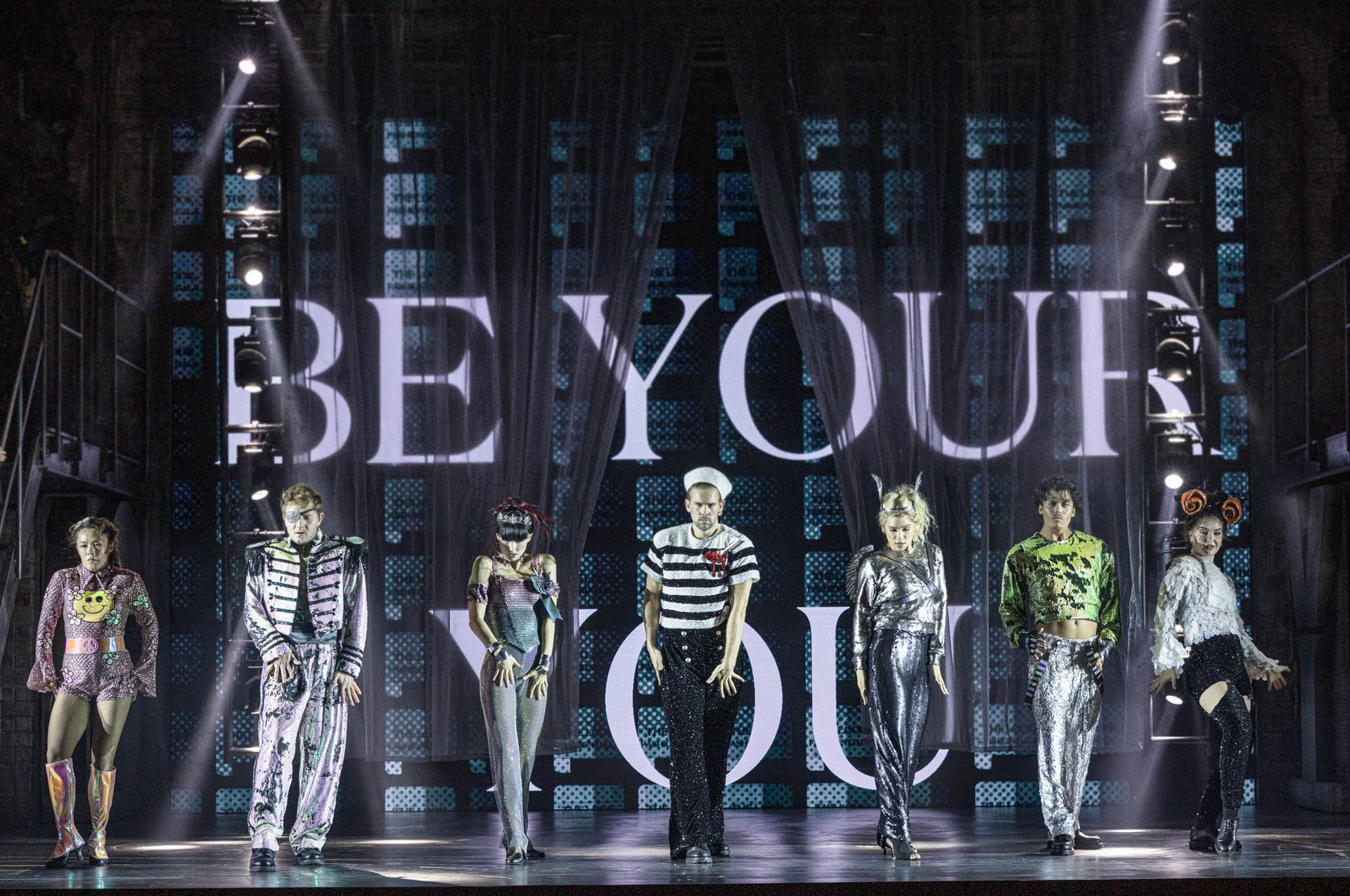 Scenbild från musikalen Joyride med ensemblen i färgstarka kostymer framför den lysande texten "BE YOUR YOU".
