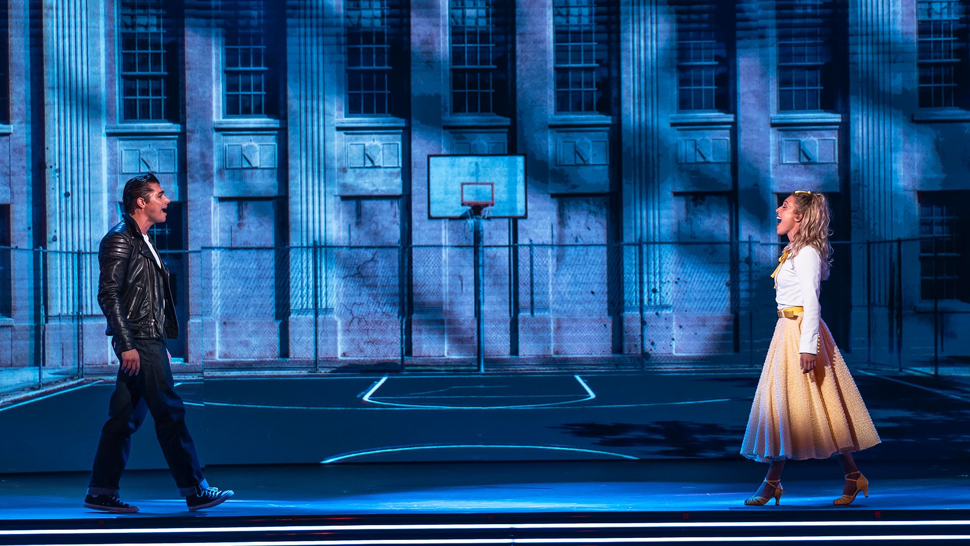 Anton Ewald som Danny och Lovisa Bengtsson som Sandy i en scen ur Grease The Musical på Rondo i Göteborg, framför en blåbelyst skolgård.