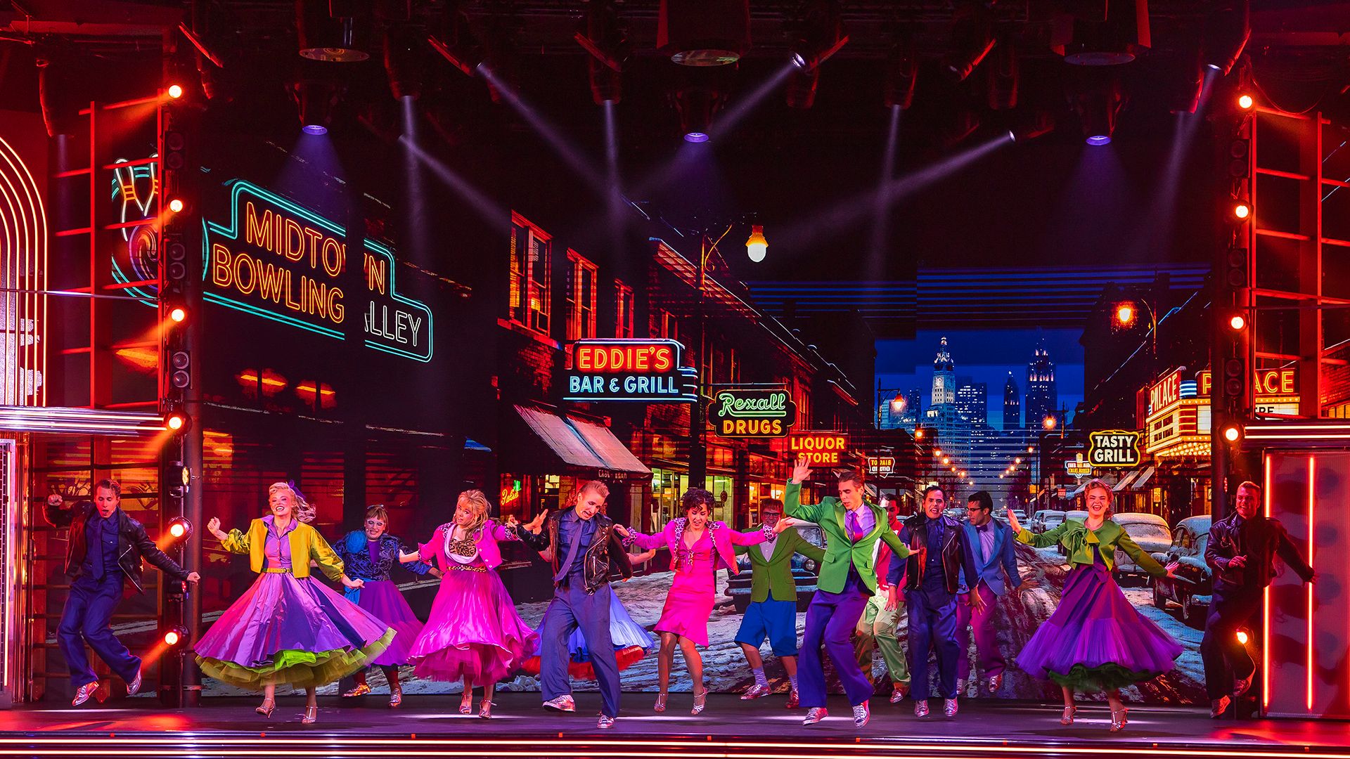 Ensemble från Grease The Musical på Rondo i Göteborg dansar på scen i färgstarka 50-talskläder, framför en neonbelyst stadsmiljö.