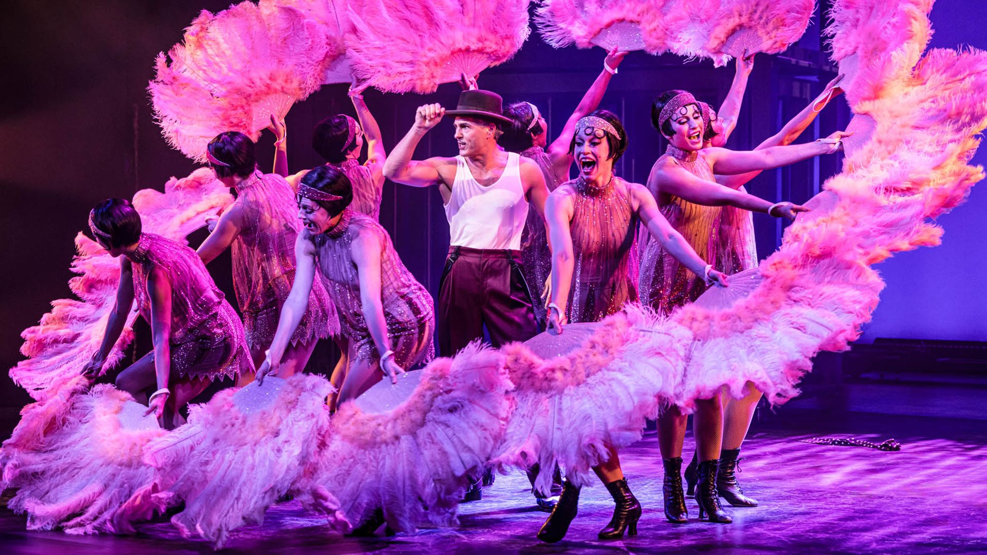 Scenbild från musikalen Chicago på Oscarsteatern i Stockholm. Peter Jöback står i mitten av ensemblen, omgiven av dansare i glittrande scenkostymer som håller stora rosa fjäderfjädrar. Scenen är upplyst i starka rosa och lila toner. Chicago spelas på Oscarsteatern 16 september–19 december.