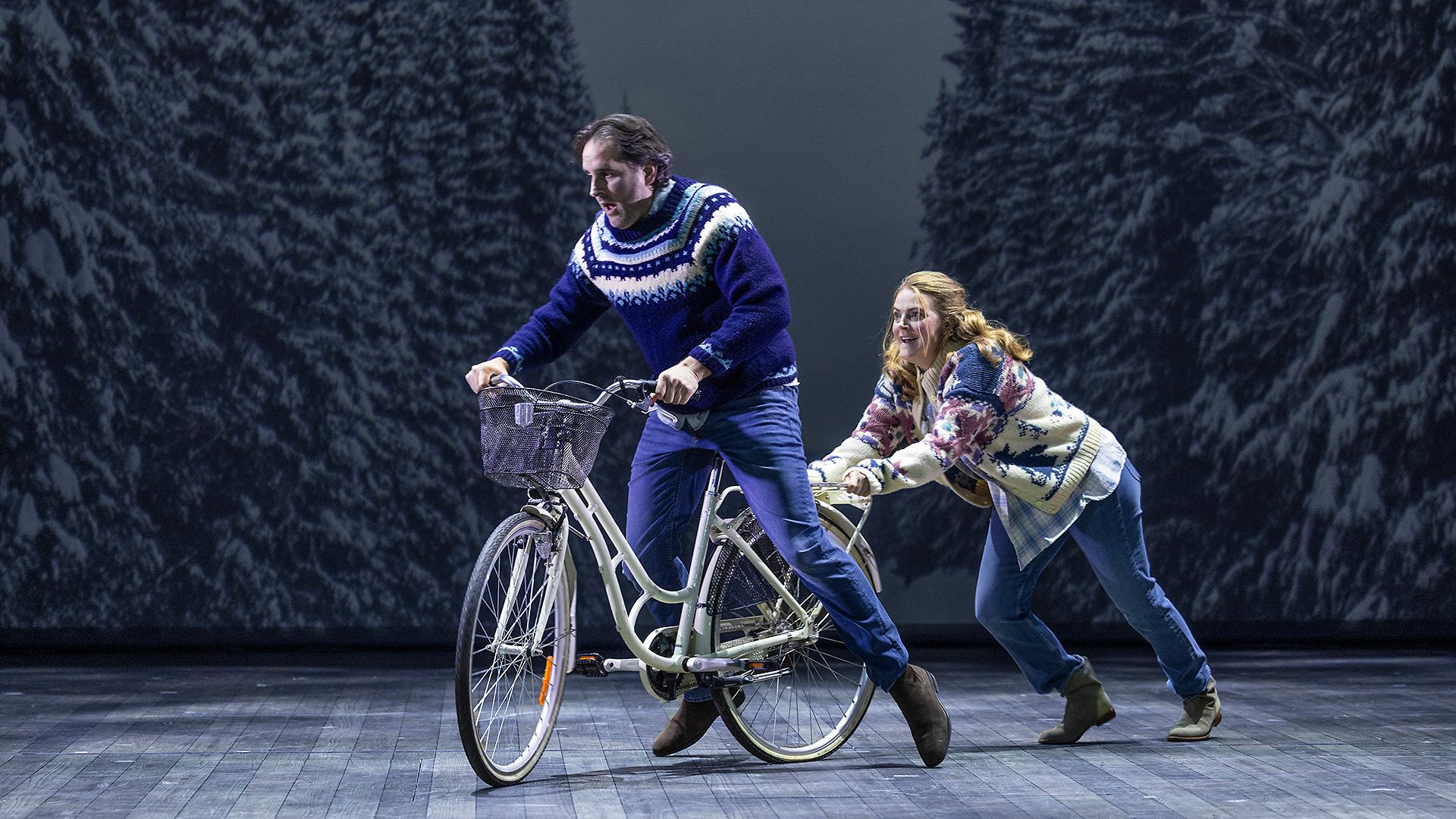 Scenbild från musikalen Så som i himmelen på Lorensbergsteatern i Göteborg. Philip Jalmelid sitter på en cykel medan Tuva Børgedotter Larsen springer bakom och skjuter på, framför en vintrig bakgrund med snötäckta granar.