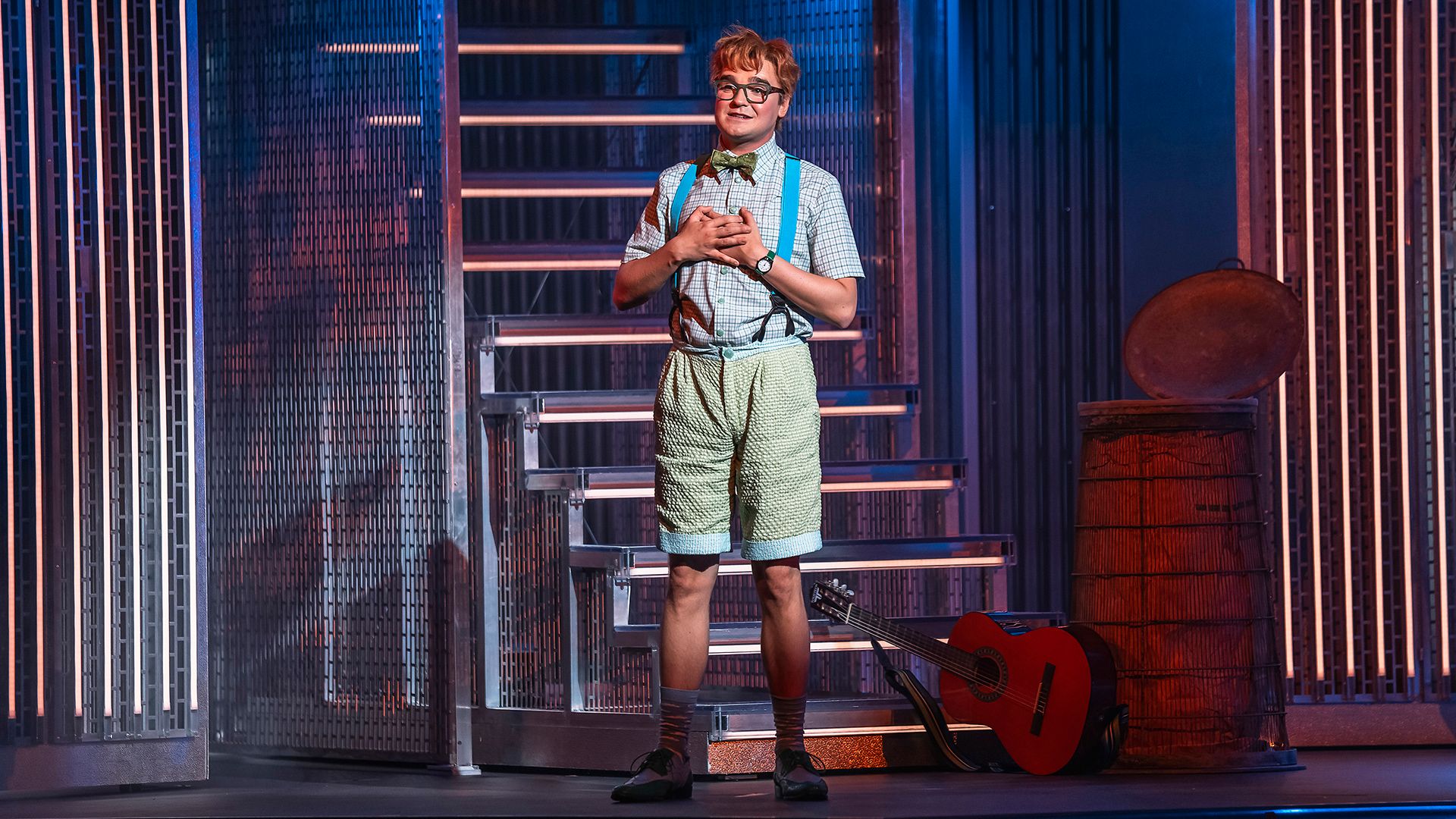 Emil Henrohn som Eugene i en scen ur Grease The Musical på Rondo i Göteborg. Han står vid en trappa i glasögon, fluga och hängslen, med en röd gitarr bredvid sig.