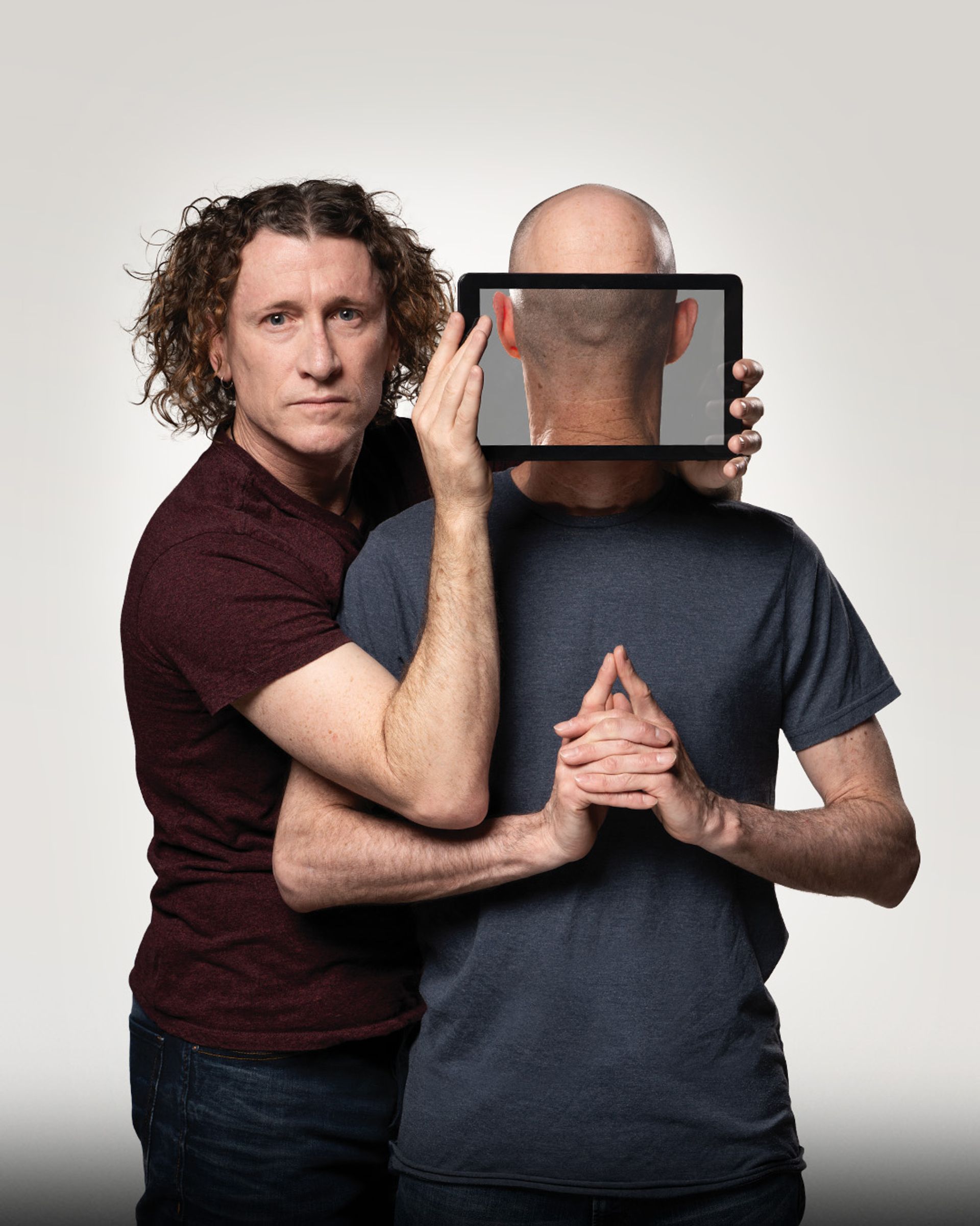 Manér bild för The Umbilical Brothers - The Distraction. The Umbilical Brothers står båda med kroppen mot kameran men den ena brodern håller en iPad framför den andras ansikte så det ser ut som att han står med ryggen mor kameran.