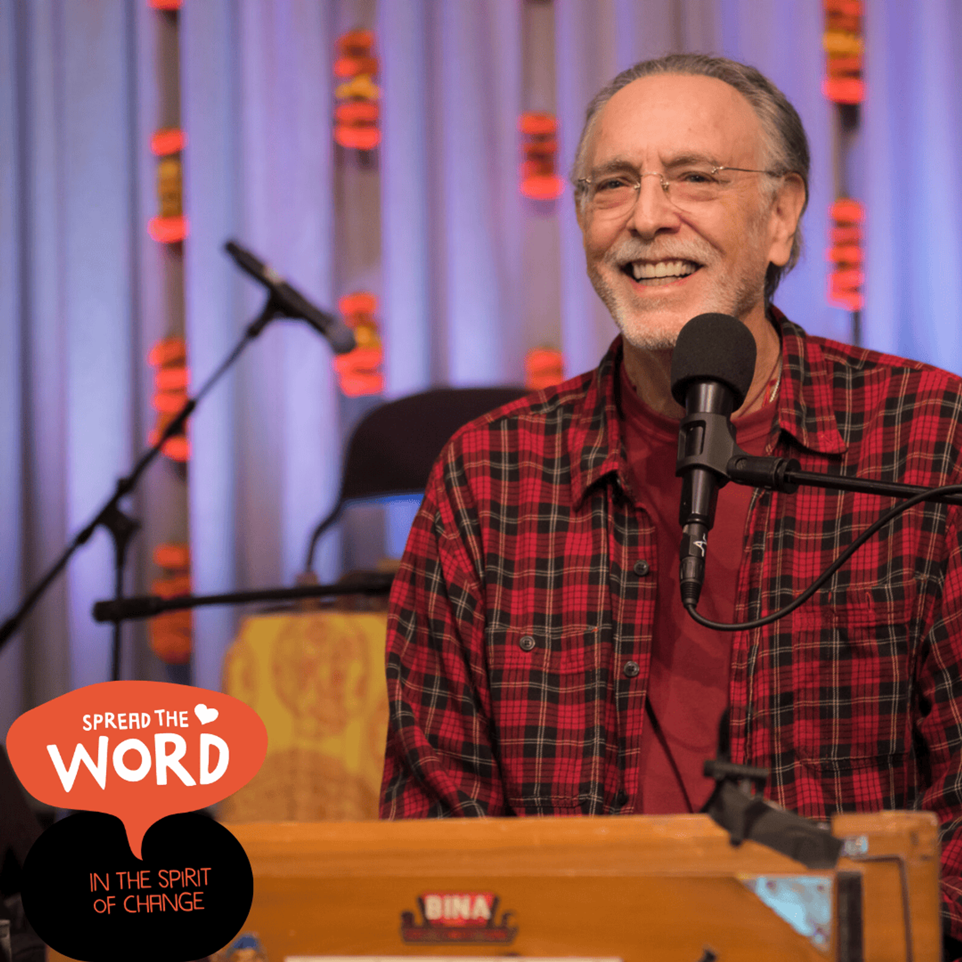 Manér bild för föreställningen Kirtan med Krishna Das. Krishna står vid ett podium med en mikrofon framför dig. Bilden är dagen på nära håll. Han har på sig en röd rutig skjorta.