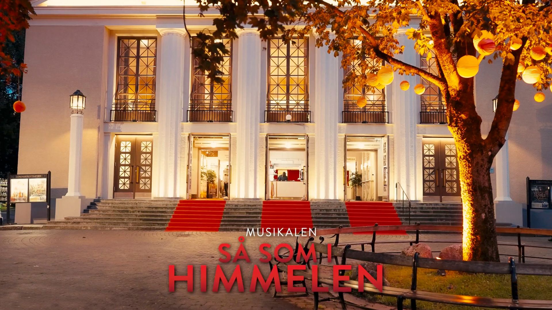 Exteriörbild av Lorensbergsteatern i Göteborg under premiärkvällen för musikalen Så som i himmelen. Teaterns upplysta entré har röda mattor och omges av höstträd med lysande lyktor. Text i bilden: ”Musikalen Så som i himmelen”.