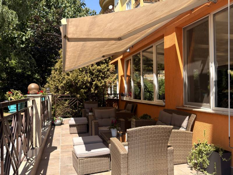 The terrace on a sunny day: an awning over wicker sofas and stools, the building’s orange façade and blue sky.