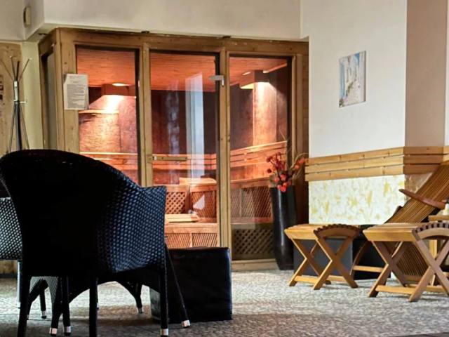 Спа зоната: плетени кресла и дървени шезлонги пред остъклената врата на сауната, вътре свети топла светлина.