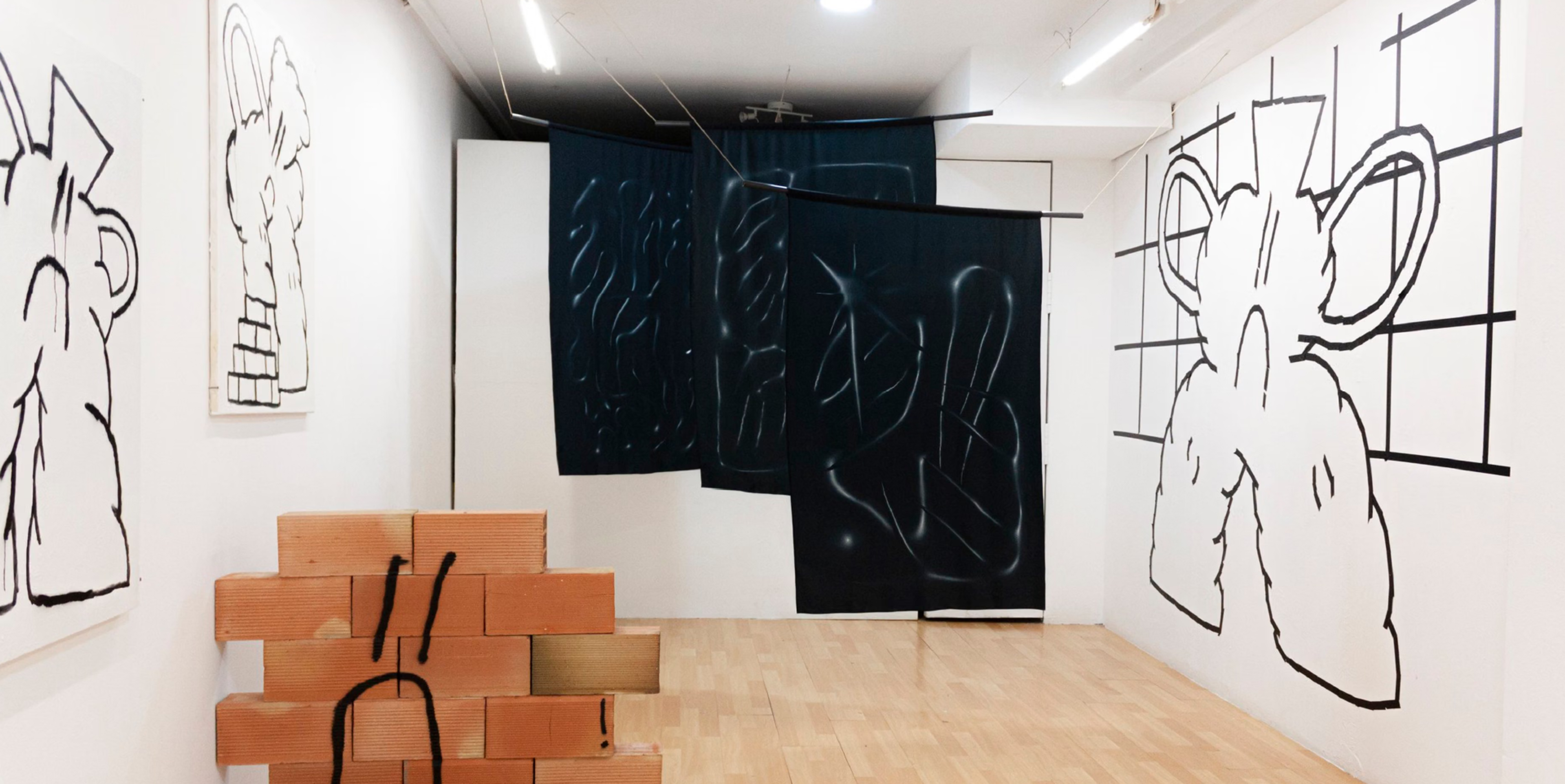 Installation view of Cinco Ceros with black line drawings, dark hanging panels and a stack of bricks.