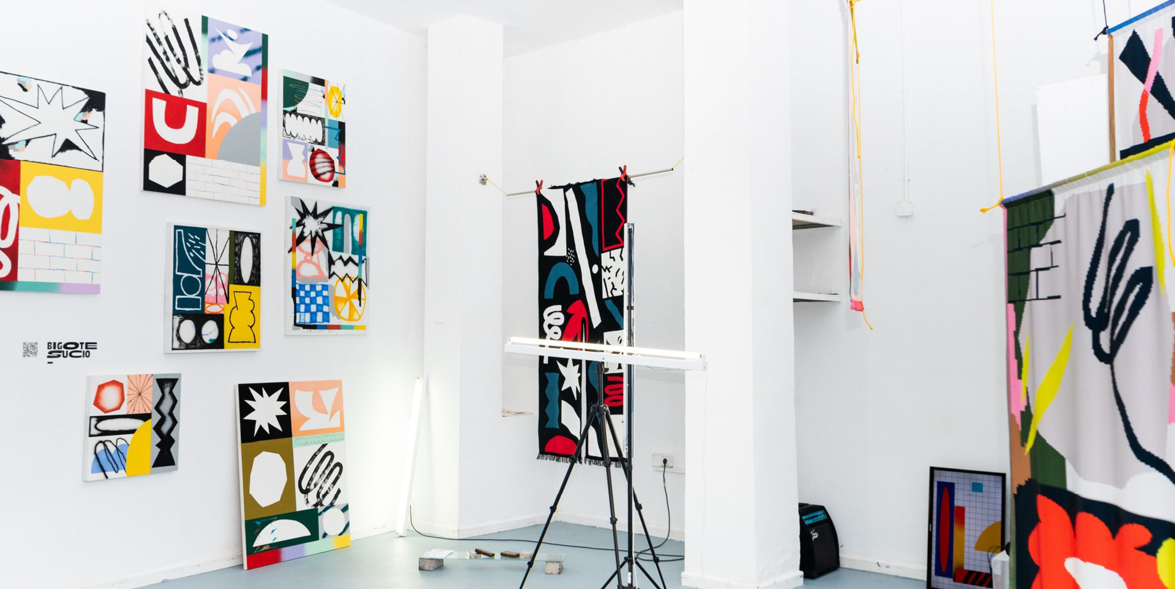 Installation view of Algoritmo Matemático Pinturesco with geometric paintings and printed fabric in a white gallery.