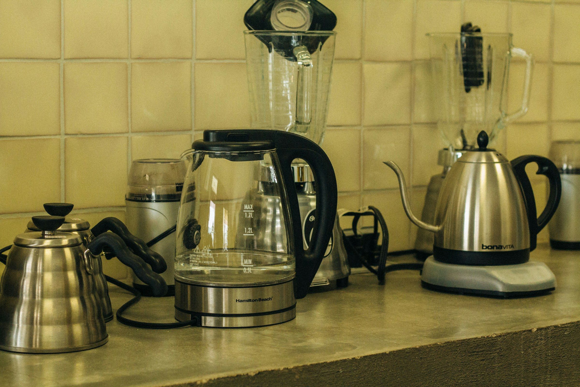 Several electric and gooseneck kettles lined up on a tiled kitchen counter