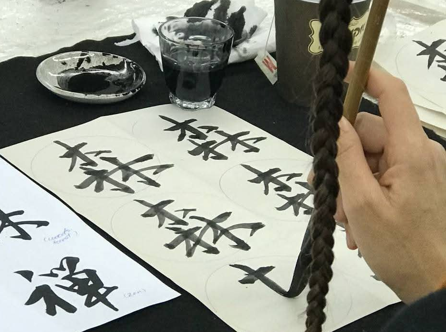 A hand holding a bamboo calligraphy brush over practice sheets of ink characters, beside an inkstone and a glass of ink