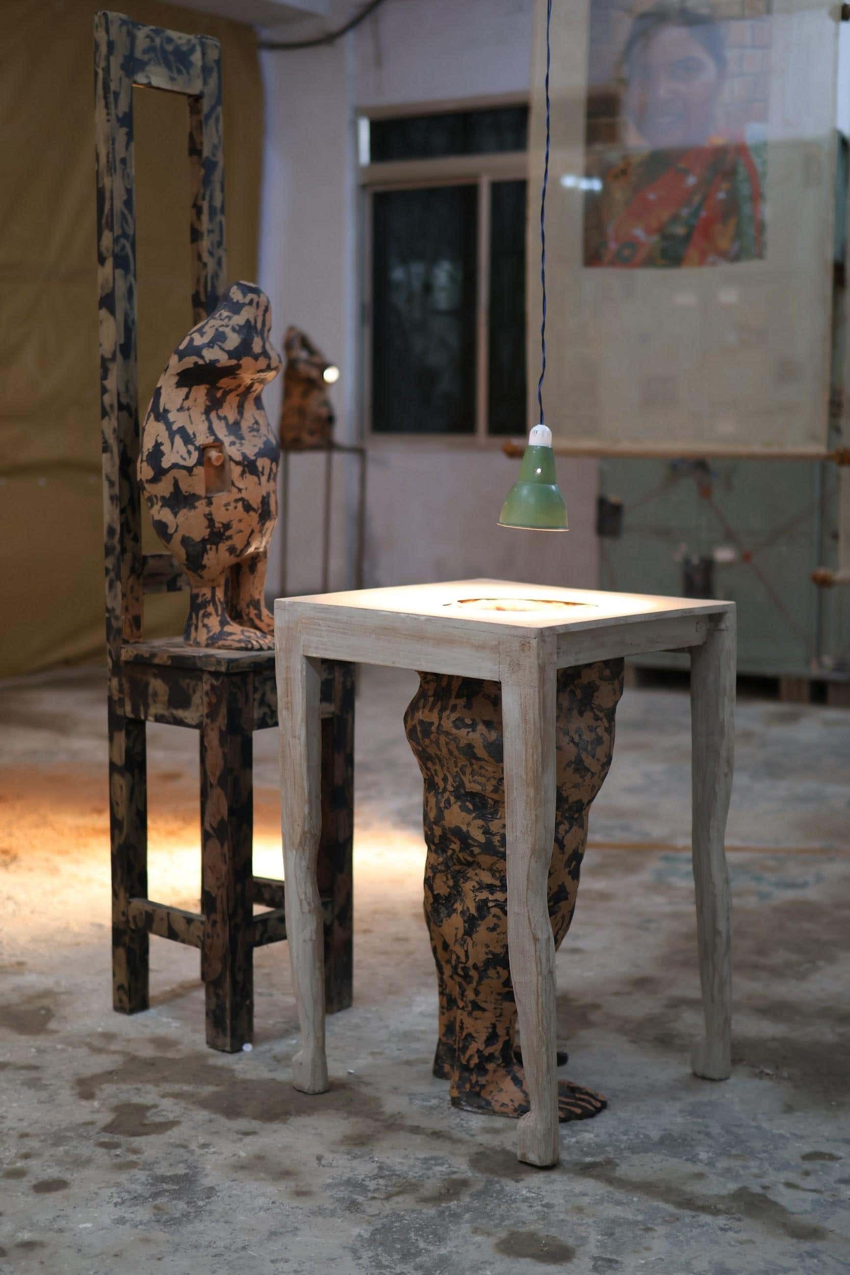A mottled terracotta figure seated on a tall-backed wooden chair, beside a pale wooden table with a body form emerging from beneath it