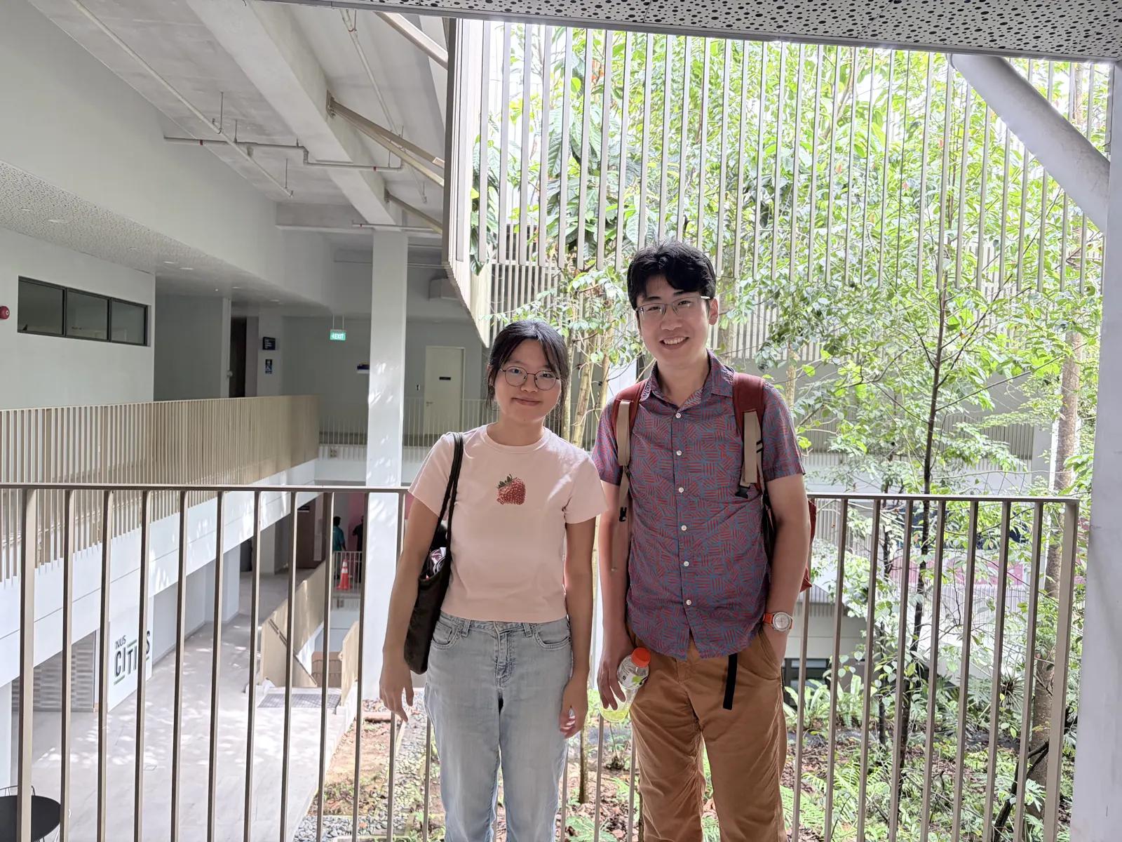 Two students smiling on an atrium walkway at the National University of Singapore, greenery behind them