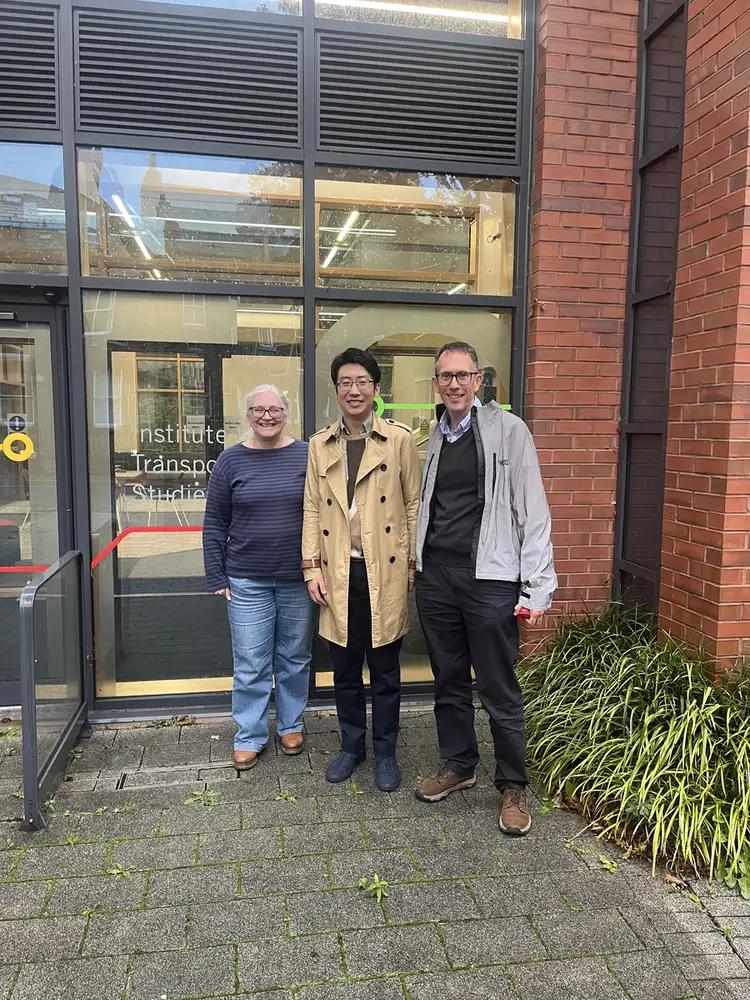 Alex in a trench coat stands with two colleagues outside the glass entrance of the Institute for Transport Studies.