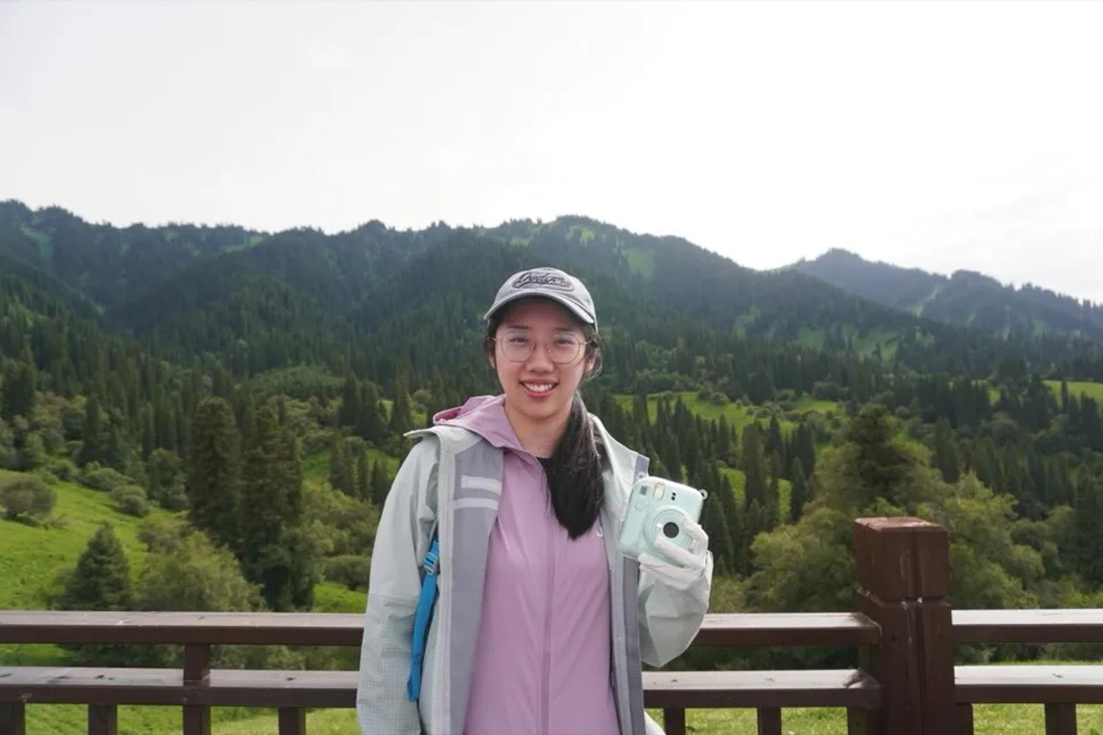 Tianle Luo holding an instant camera in front of forested mountains