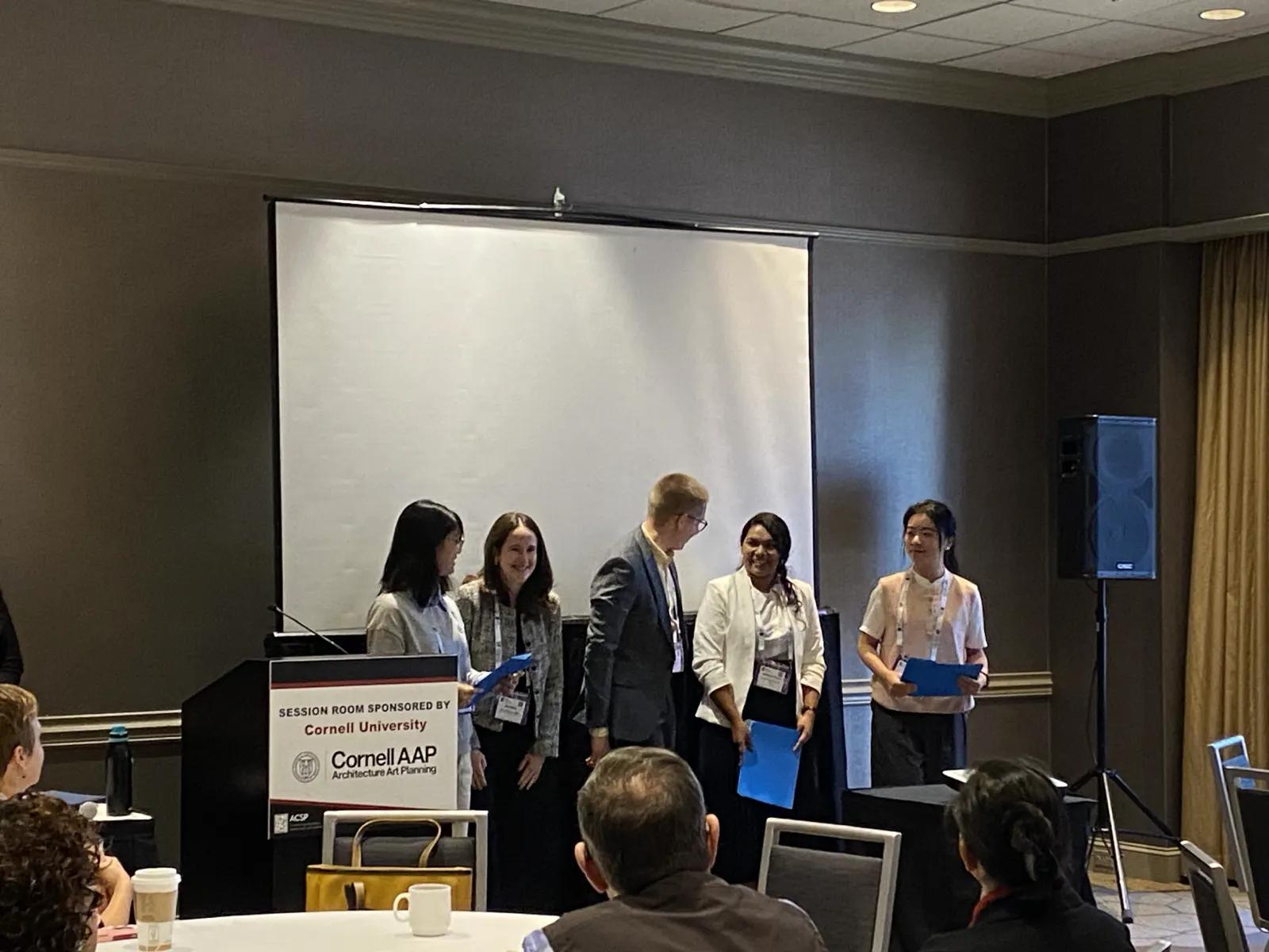 Five people standing on stage at a conference session, three award-winning students with the two co-chairs of the case study selection committee