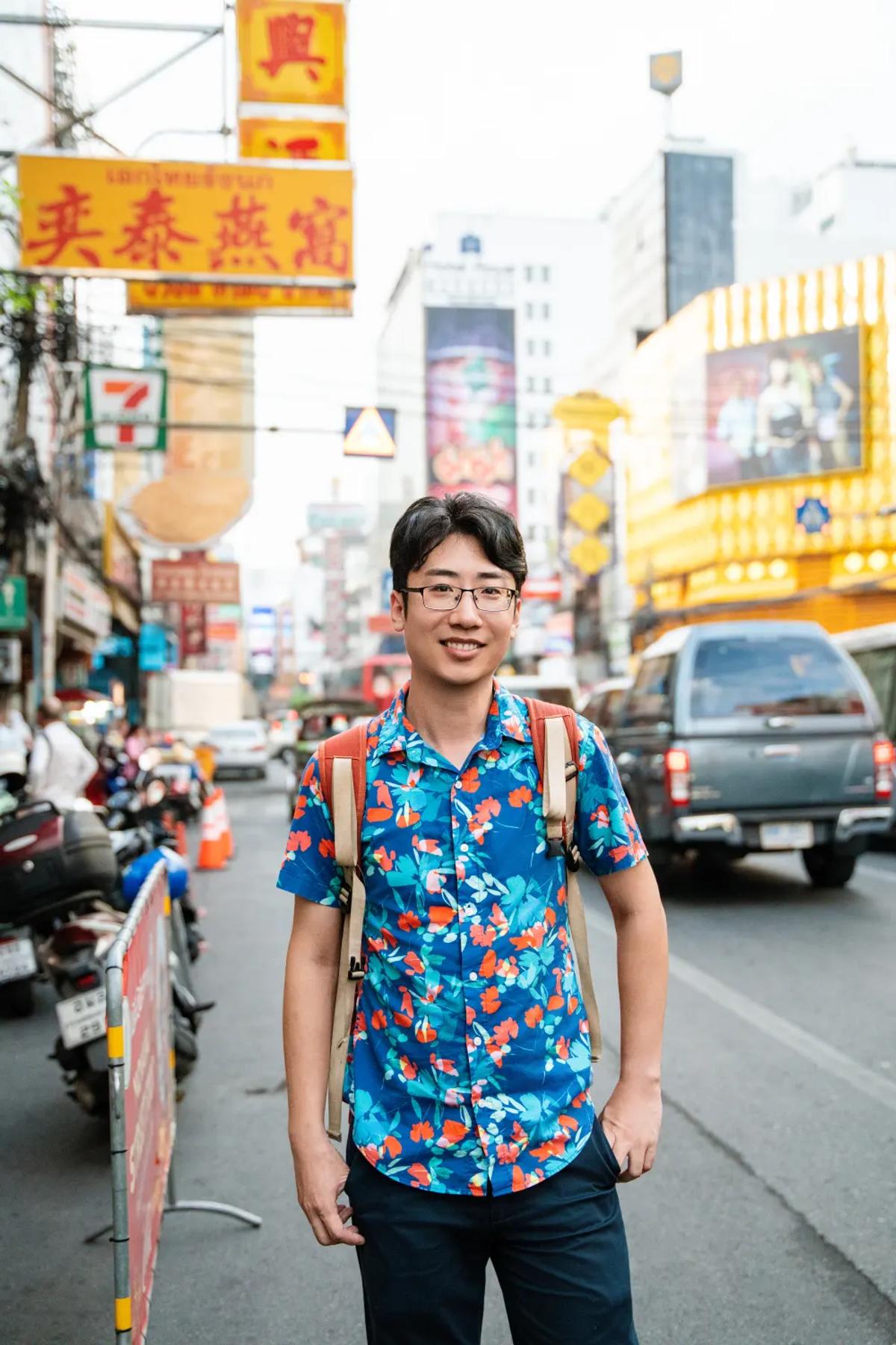 Shengxiao (Alex) Li smiling on a busy street in Chinatown, Bangkok, surrounded by shop signs and traffic