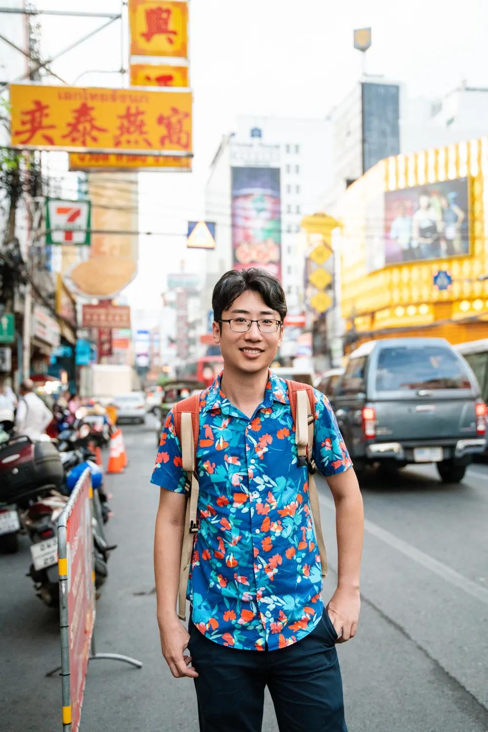 Shengxiao (Alex) Li smiling on a busy street in Chinatown, Bangkok, surrounded by shop signs and traffic