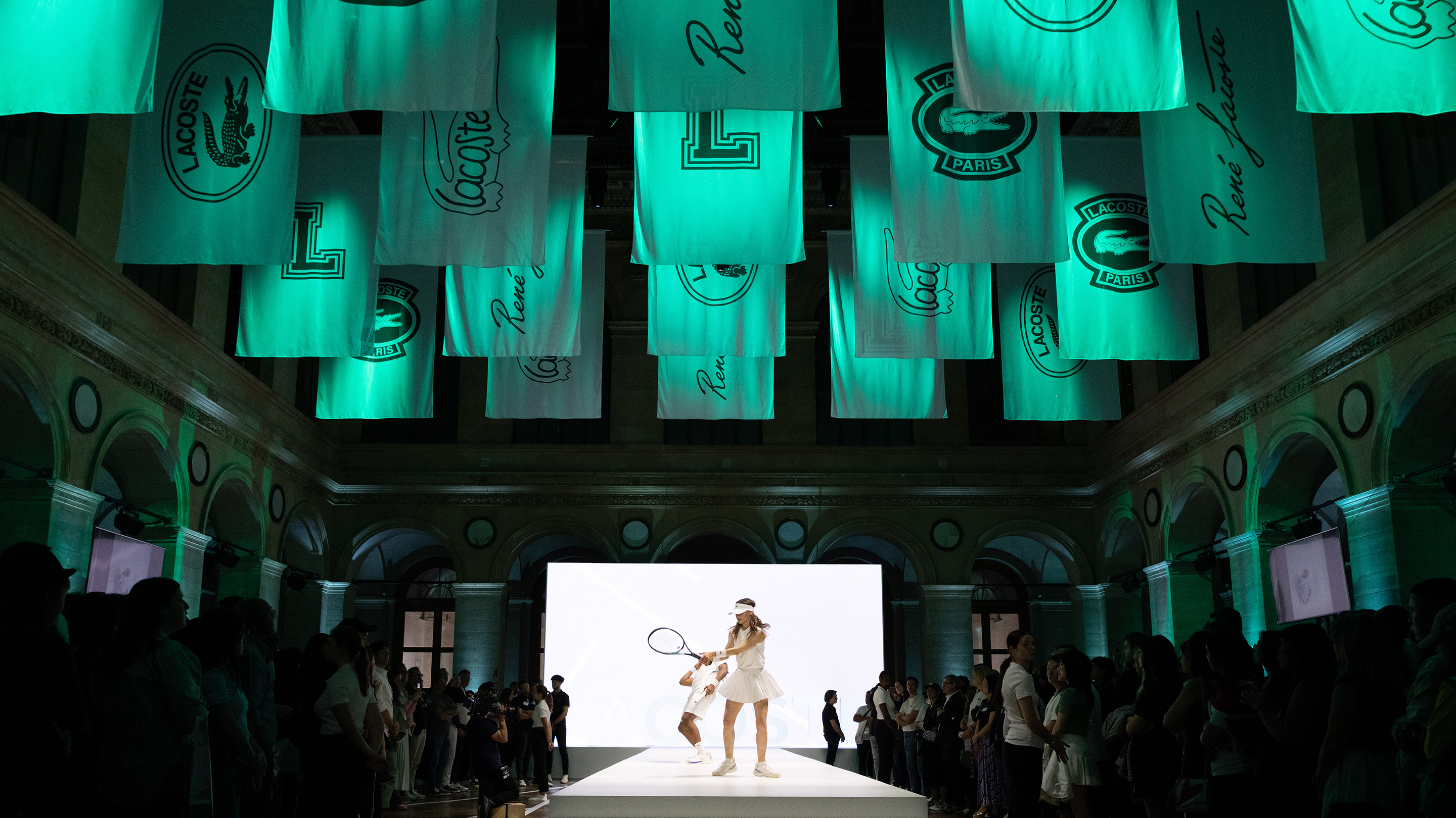 Lacoste fashion show with models walking the runway at Palais Brongniart in Paris featuring suspended green Lacoste flags across the ceiling.