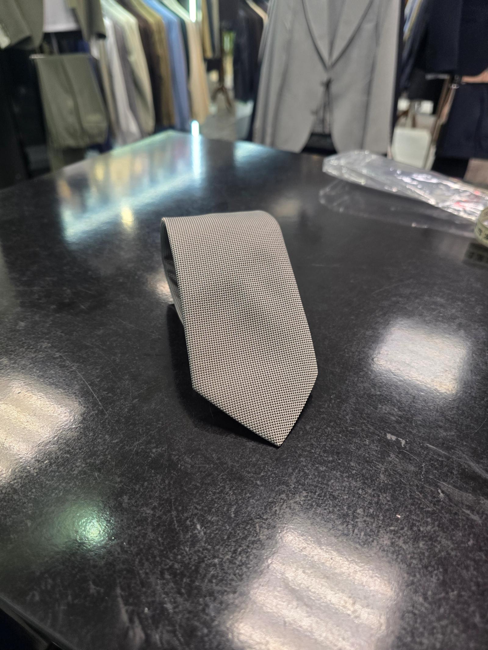 Grey slim men's tie with fine micro-texture displayed on a black table with tailoring in the background.