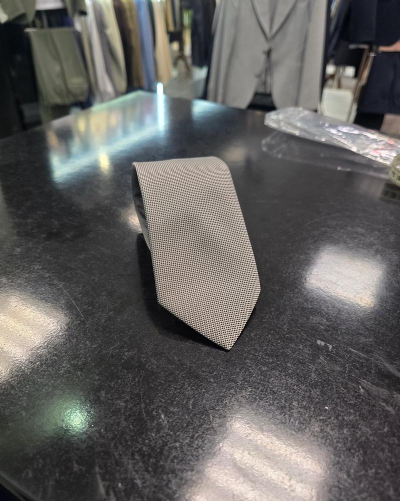 Grey slim men's tie with fine micro-texture displayed on a black table with tailoring in the background.