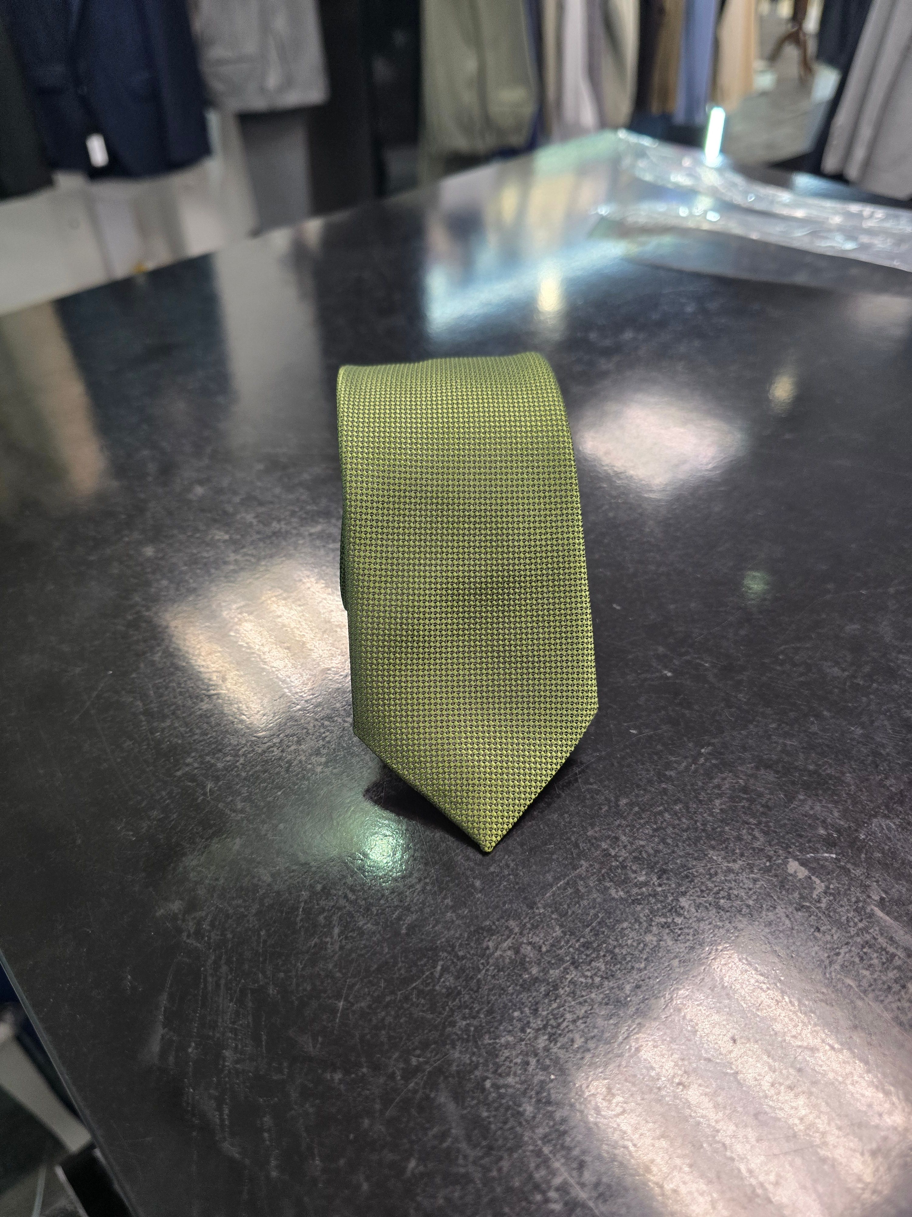 Olive slim tie with fine micro-texture rolled neatly on a black table with tailoring in the background