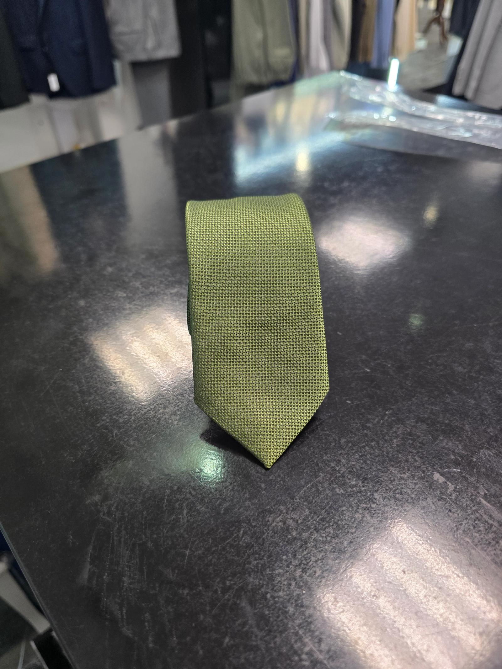 Olive slim tie with fine micro-texture rolled neatly on a black table with tailoring in the background