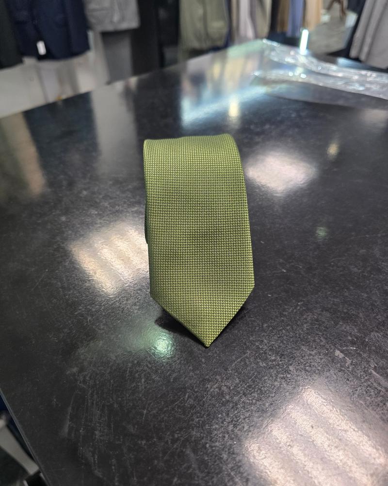 Olive slim tie with fine micro-texture rolled neatly on a black table with tailoring in the background