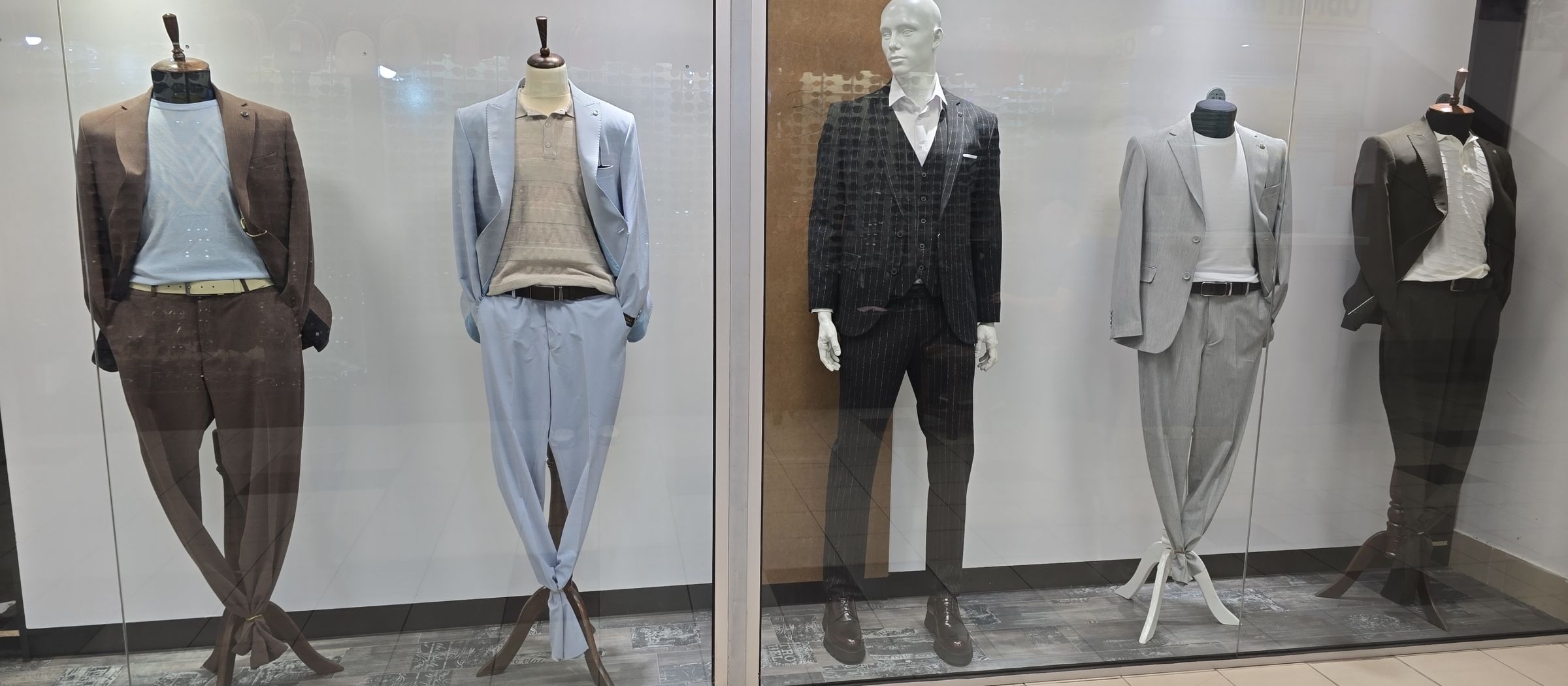 A storefront display featuring five mannequins dressed in formal and semi-formal men's attire. From left to right: a brown suit with a light blue shirt, a light blue suit with a beige polo shirt, a black pinstripe three-piece suit with a white shirt, a light gray suit with a white shirt, and a dark brown suit with a white polo shirt. The mannequins are posed against a glass window with a tiled floor visible in the foreground.