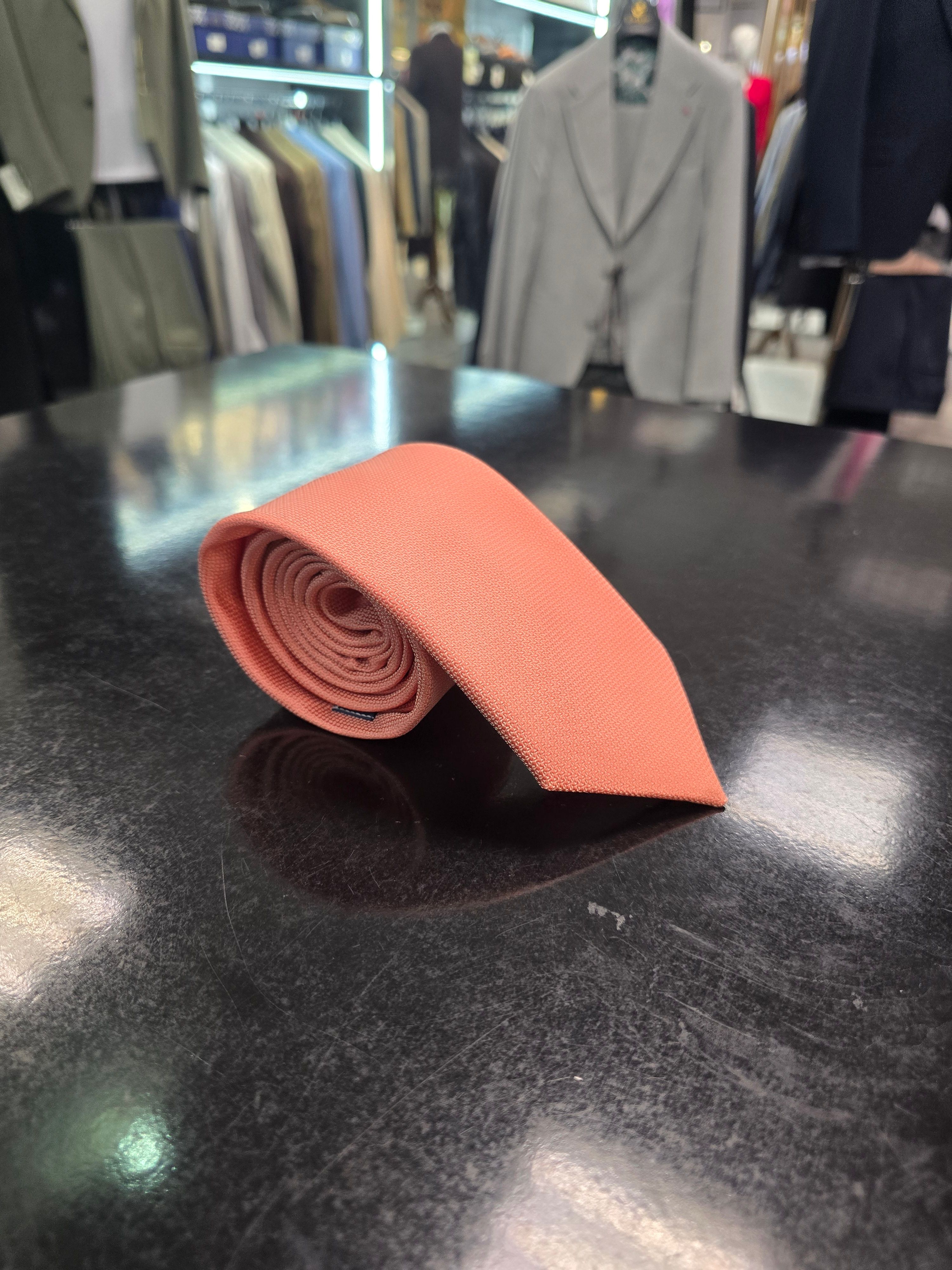 A rolled coral silk tie and matching pocket square set, displayed on a dark surface.