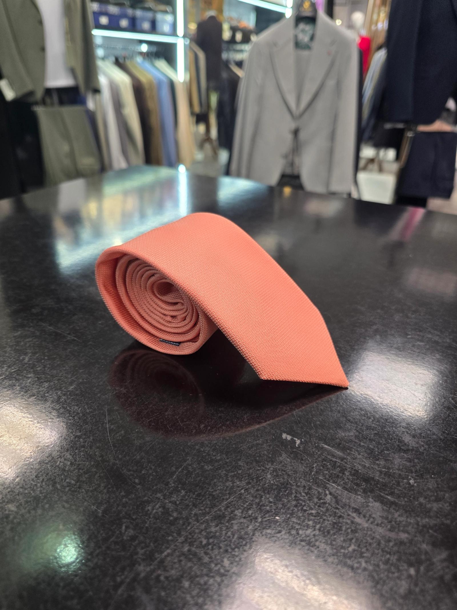 A rolled coral silk tie and matching pocket square set, displayed on a dark surface.
