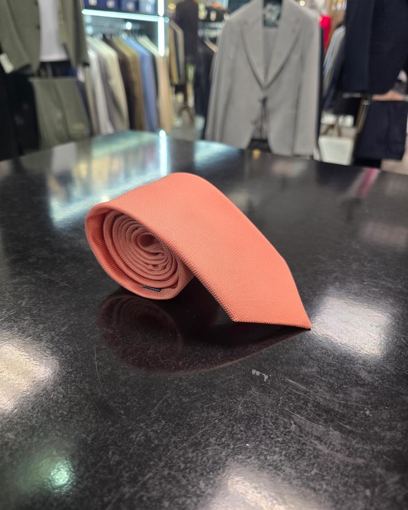 A rolled coral silk tie and matching pocket square set, displayed on a dark surface.