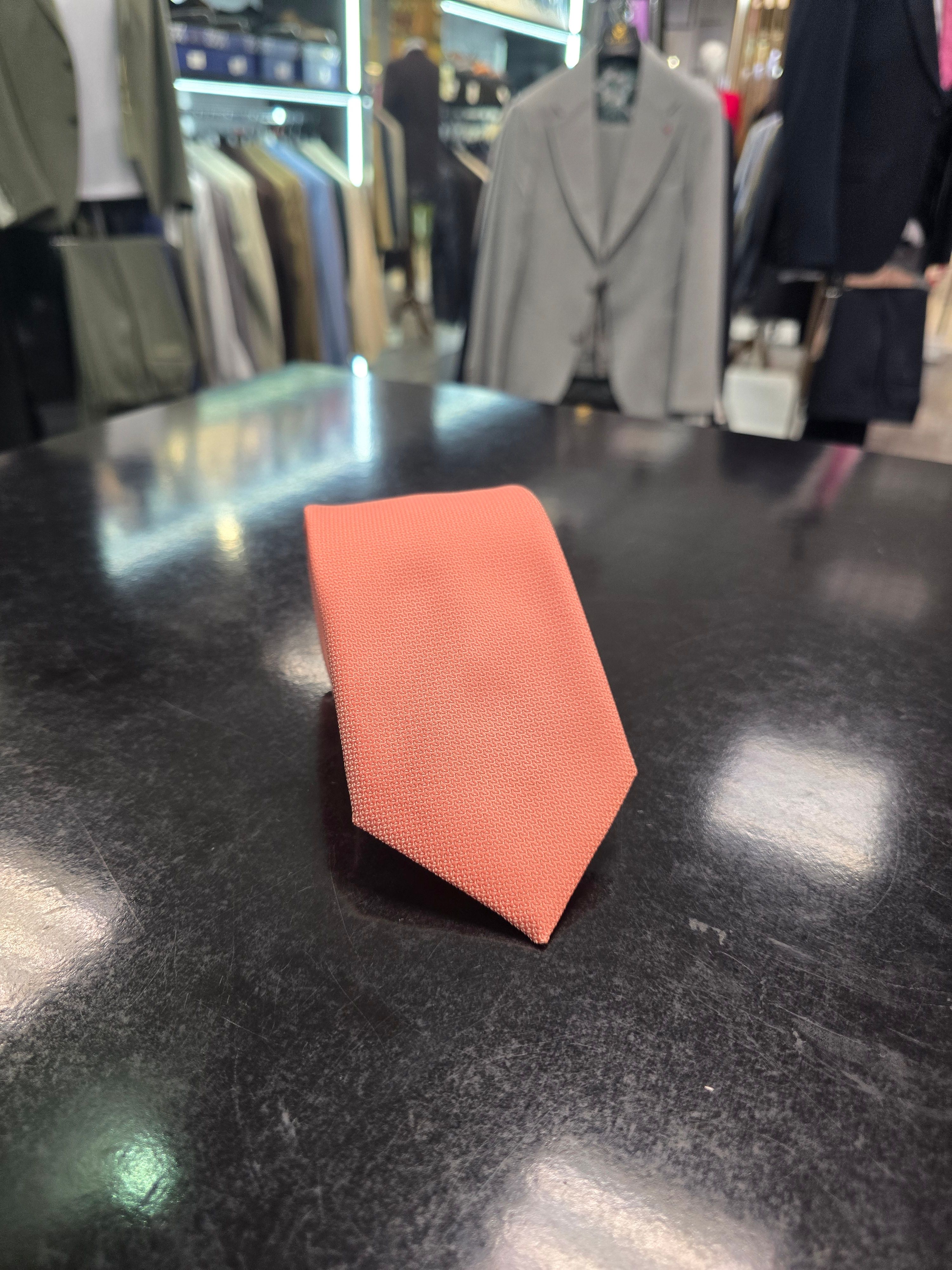 A rolled coral silk tie and matching pocket square set, displayed on a dark surface.