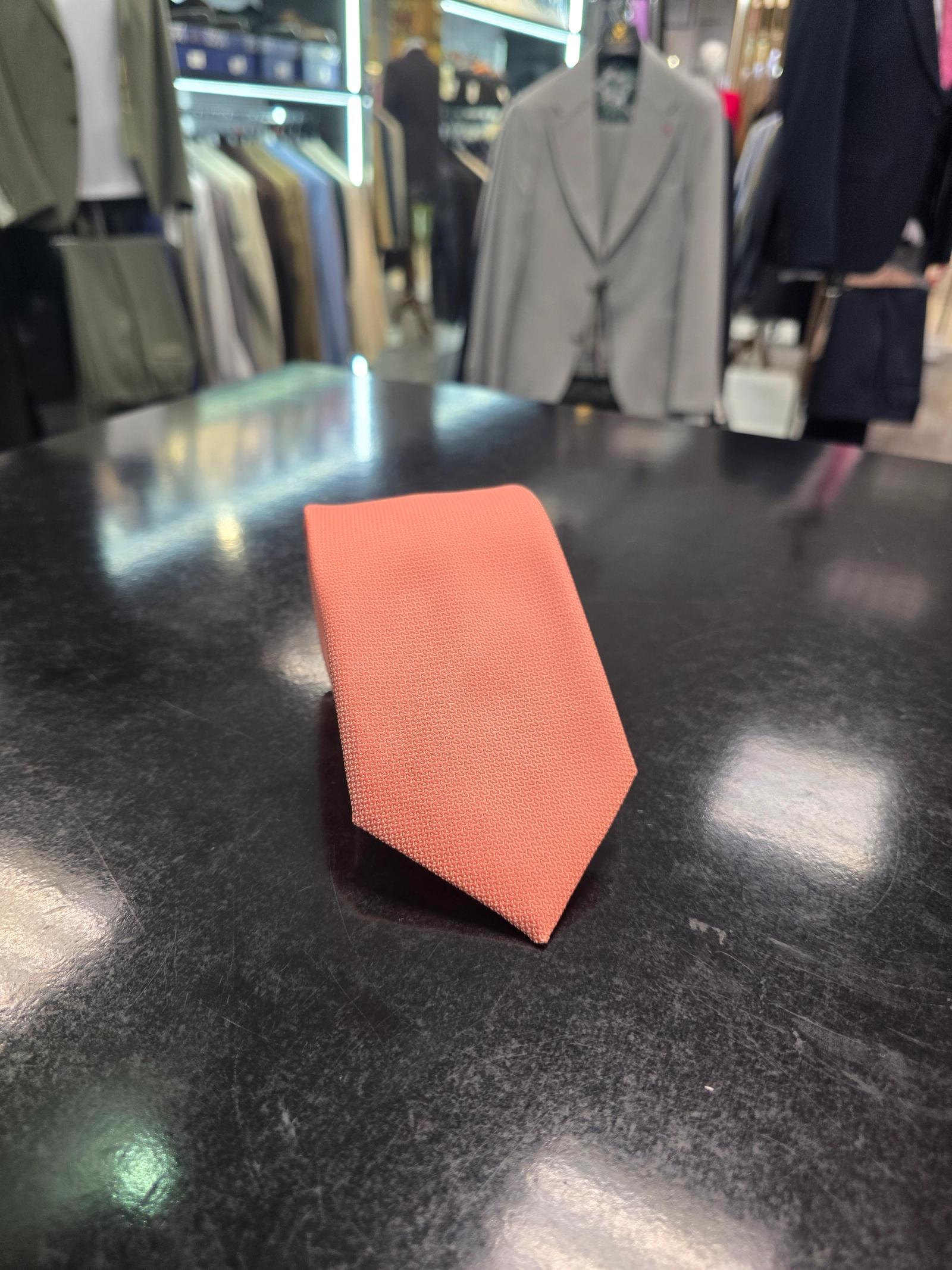 A rolled coral silk tie and matching pocket square set, displayed on a dark surface.