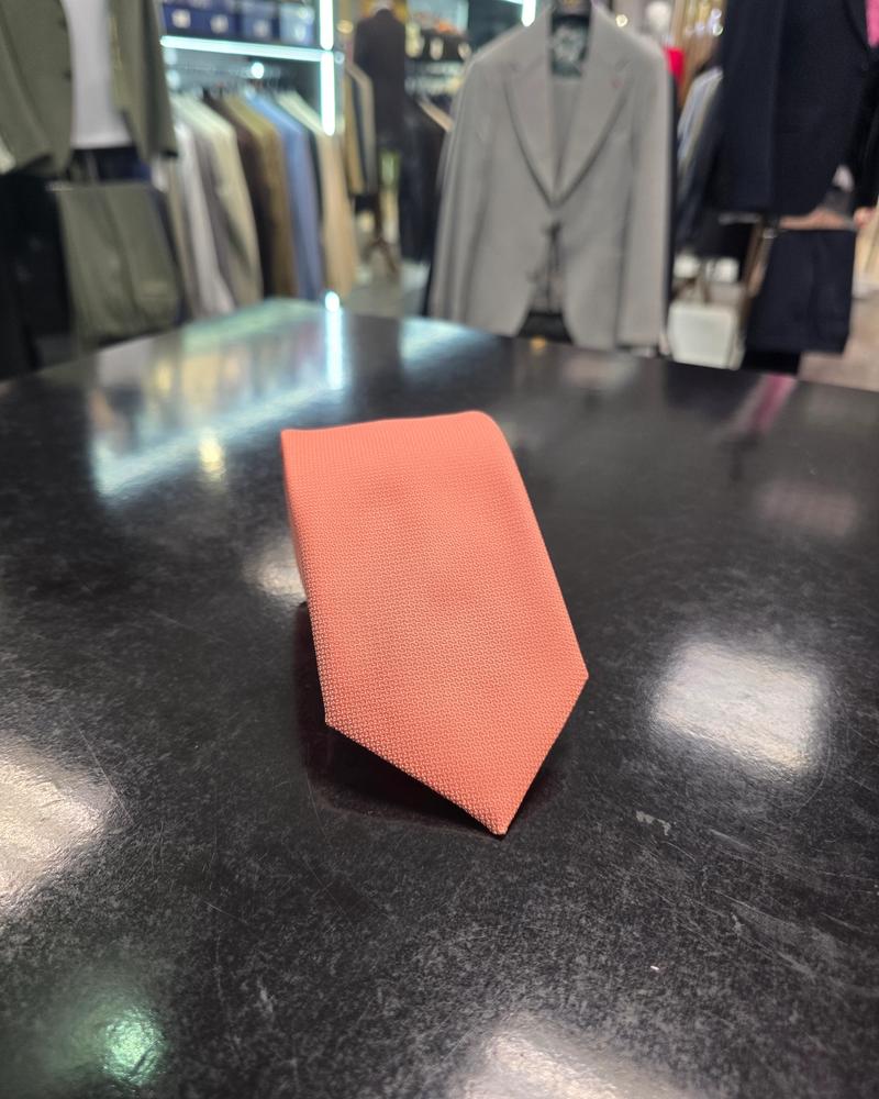A rolled coral silk tie and matching pocket square set, displayed on a dark surface.