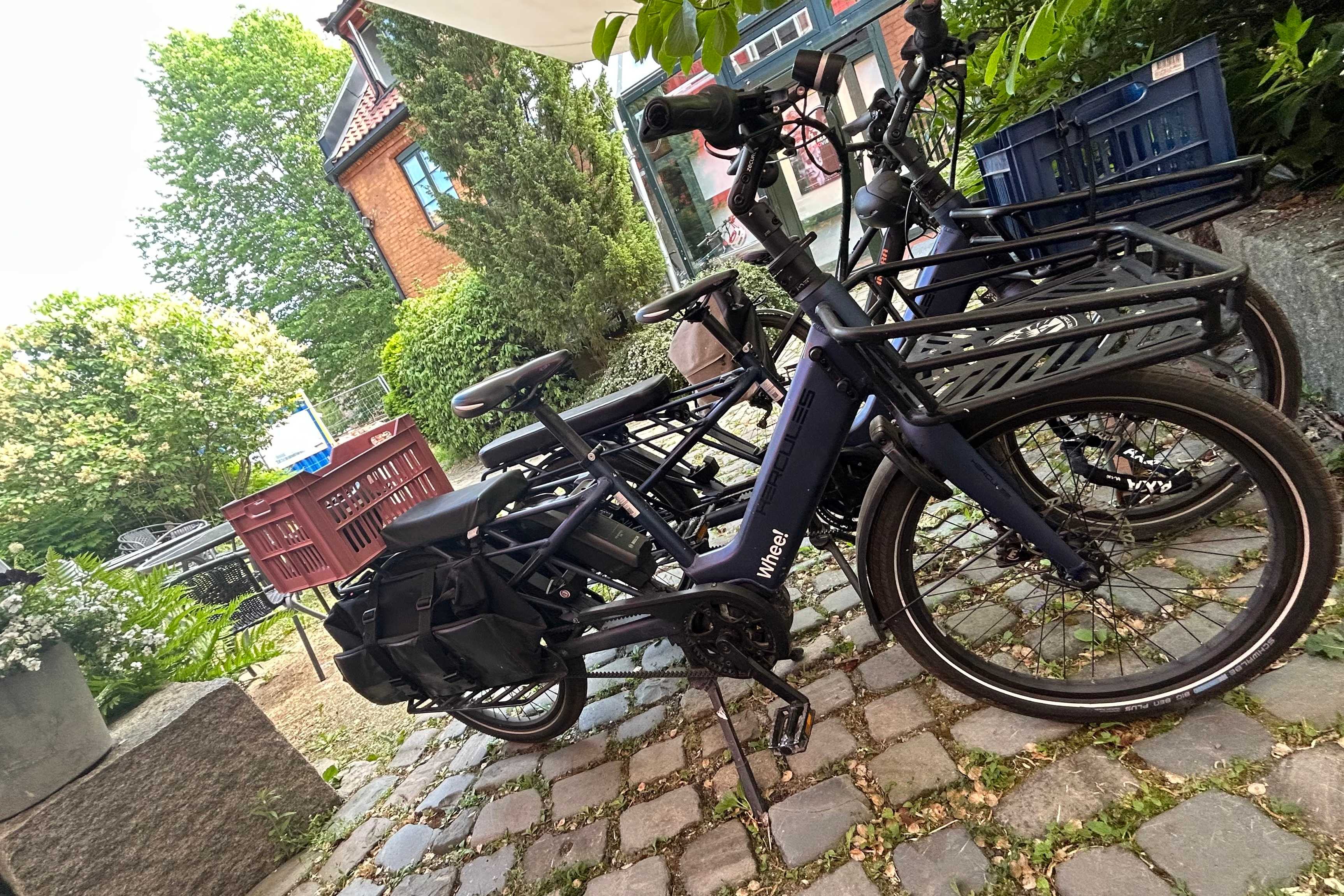 Whee!-sykler parkert ved siden av hverandre, med kurv foran og bak. De står på en hellebelagt flate, med grøntområder og noen bygninger i bakgrunnen.