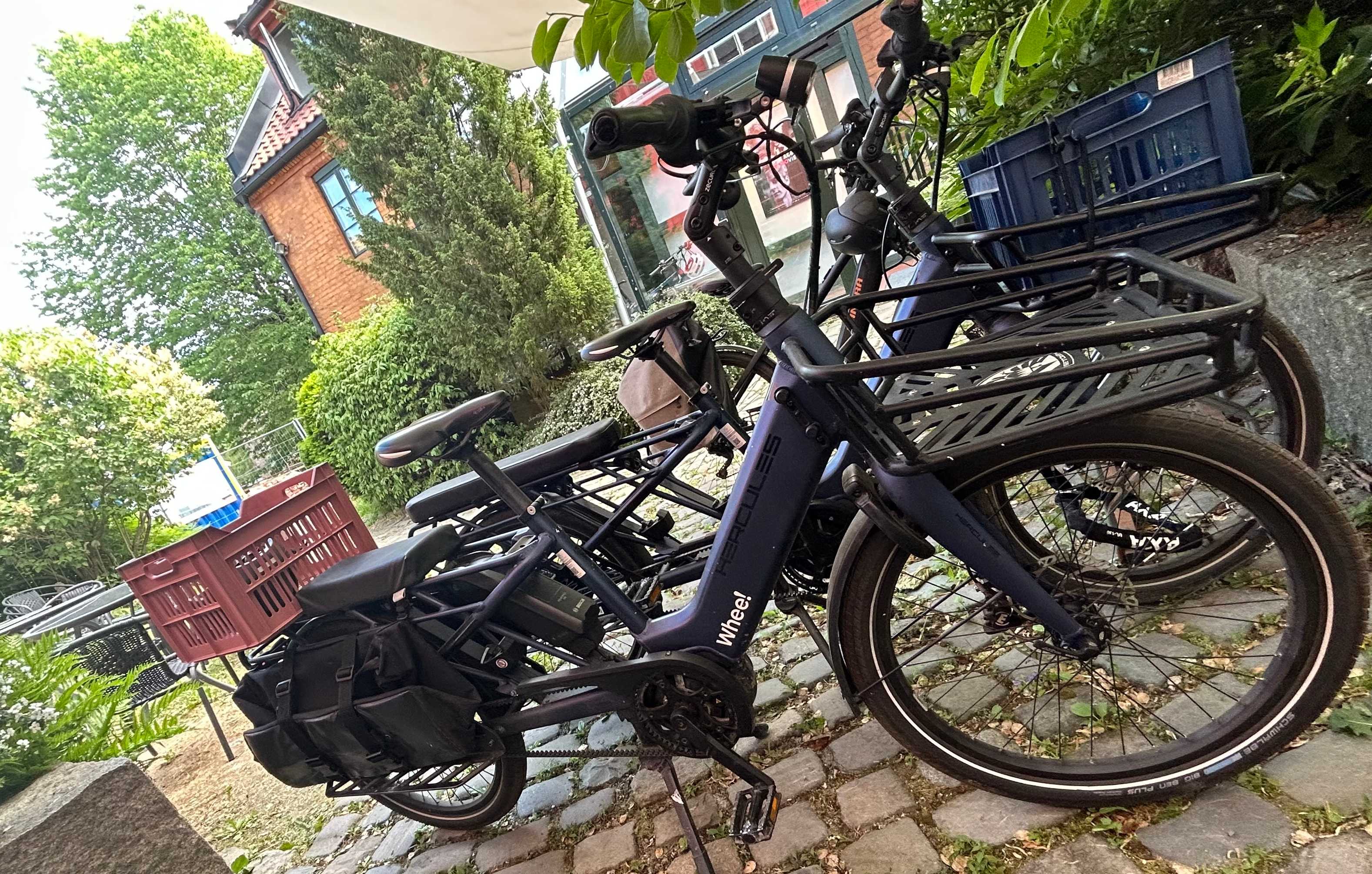 Whee!-sykler parkert ved siden av hverandre. De står på noen heller i et grøntområde. Hus og trær i bakgrunnen.