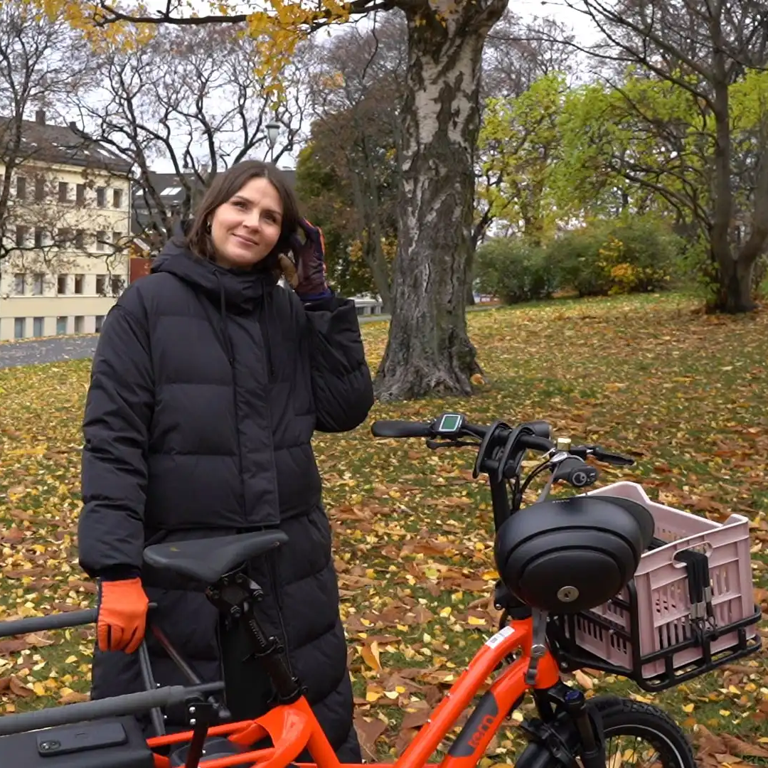Anne står bak sin Tern GSD. Hjelmen henger på styret. Hun har på seg en vinterkåpe og det er løv på bakken.