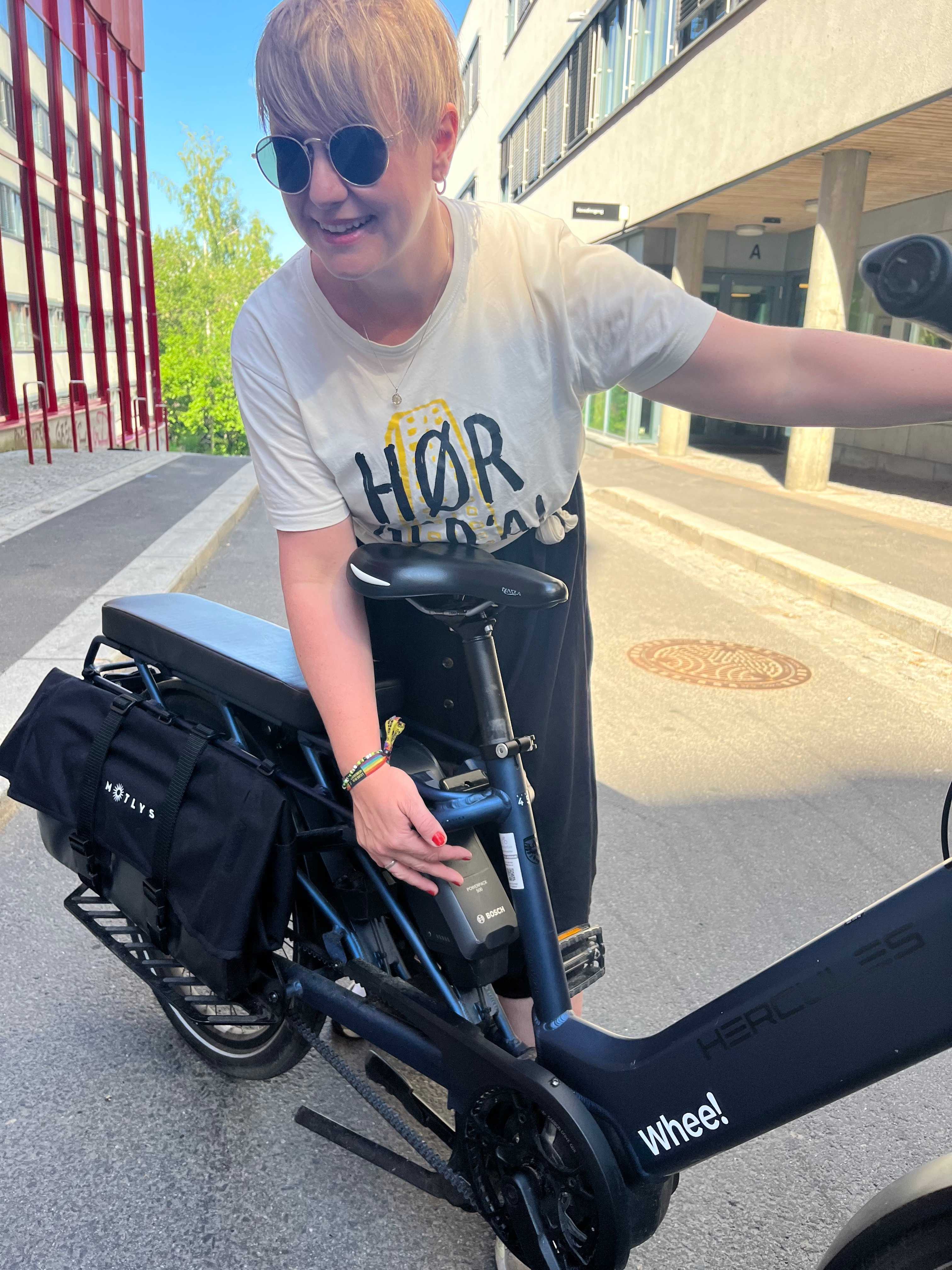 Person står og holder på en Whee!-sykkel. De har på solbriller og t-skjorte, og står mellom noe som kan se ut som et kontotbygg og et parkeringshus.