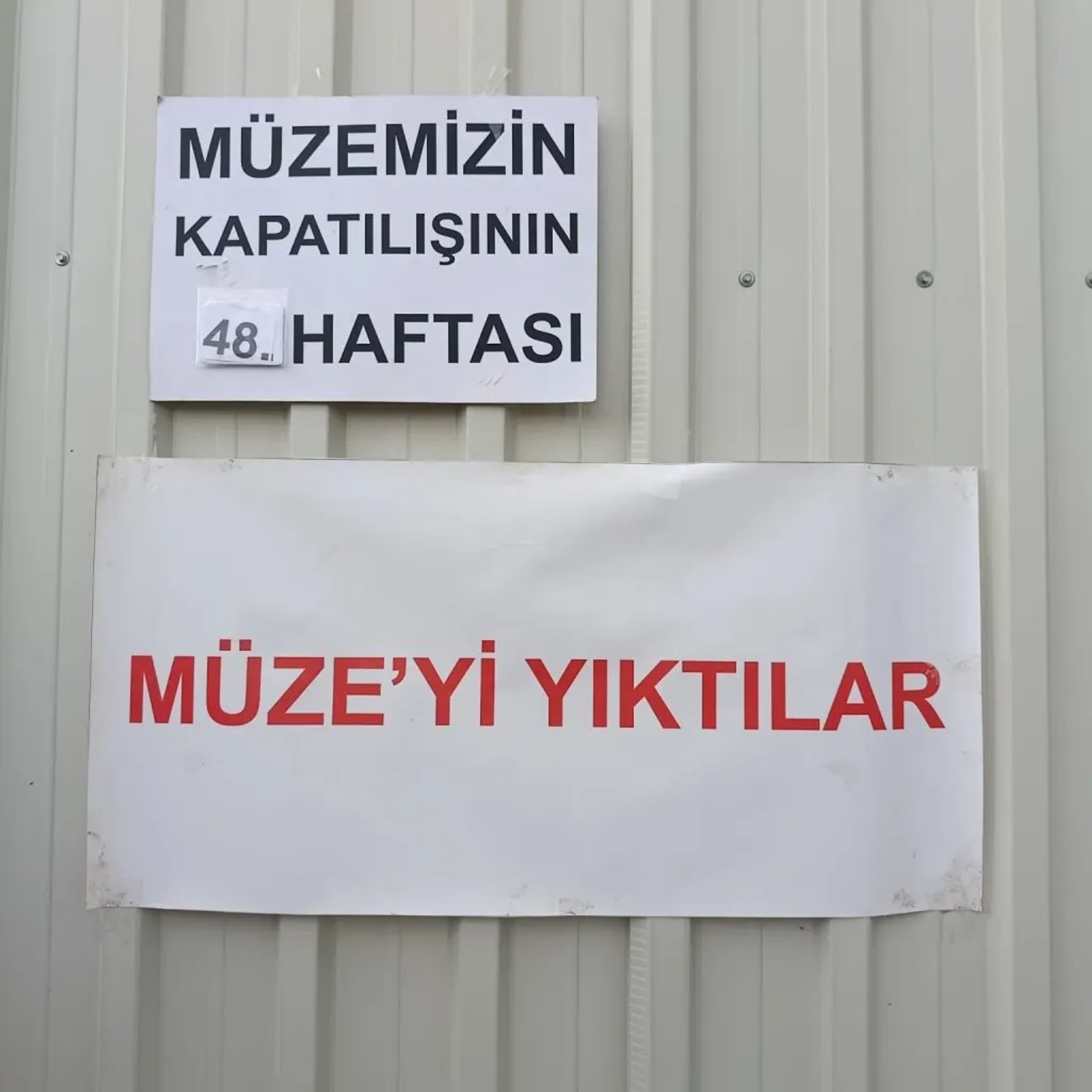 Fotoğraf betimlemesi: Görselde açık renkli, dikey oluklu metal bir yüzeye yapıştırılmış iki ayrı afiş yer alıyor. Üstteki küçük afişte büyük siyah harflerle “MÜZEMİZİN KAPATILIŞININ 48. HAFTASI” yazıyor; “48.” ifadesi sonradan eklenmiş ya da güncellenebilir biçimde ayrı bir kâğıt parçası gibi yapıştırılmış. Alt bölümde daha büyük, beyaz bir afiş bulunuyor. Üzerinde kırmızı, kalın harflerle “MÜZE’Yİ YIKTILAR” yazıyor.