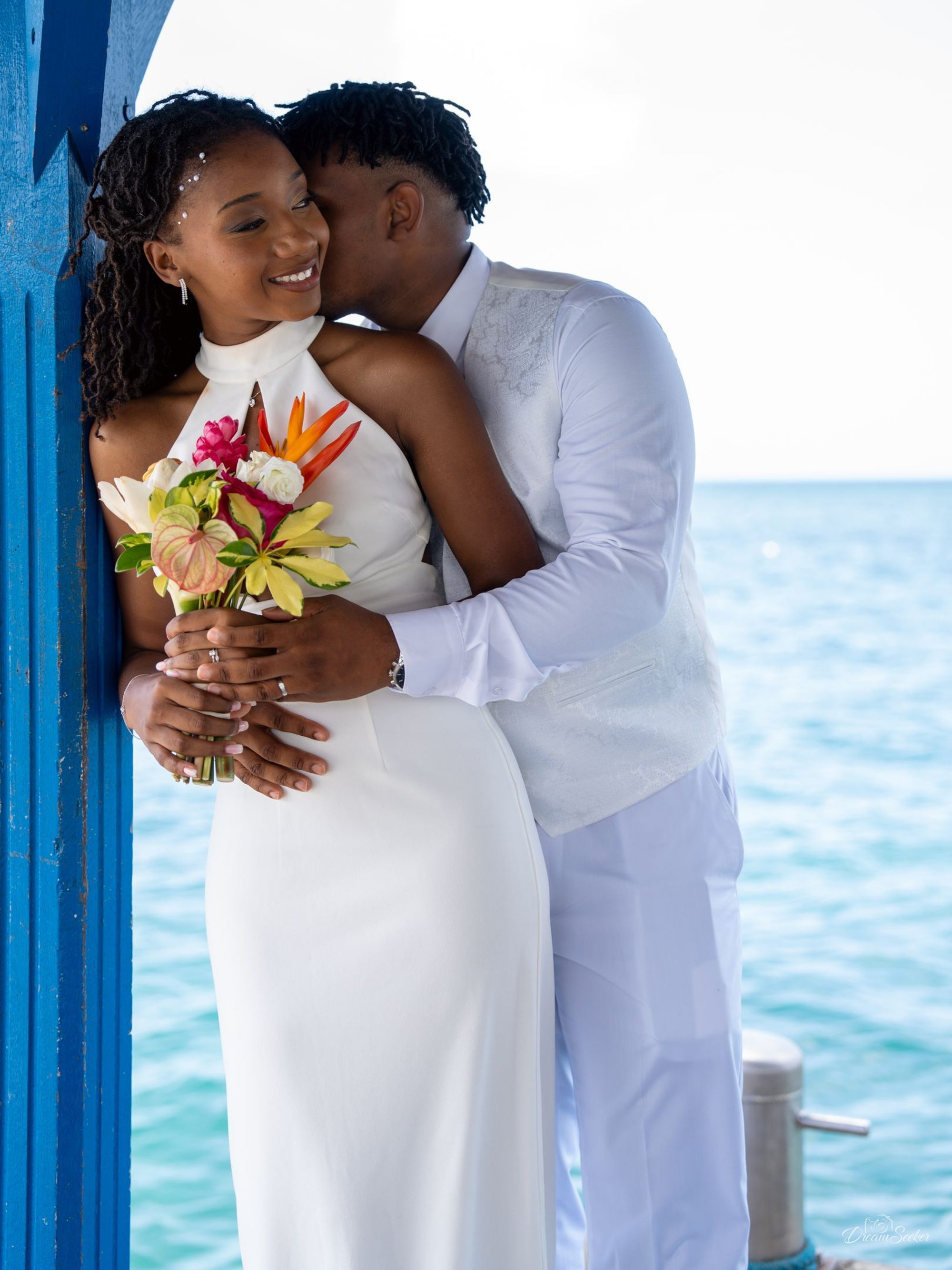 Marié embrassant la joue de la mariée devant une porte bleue, mer derrière
