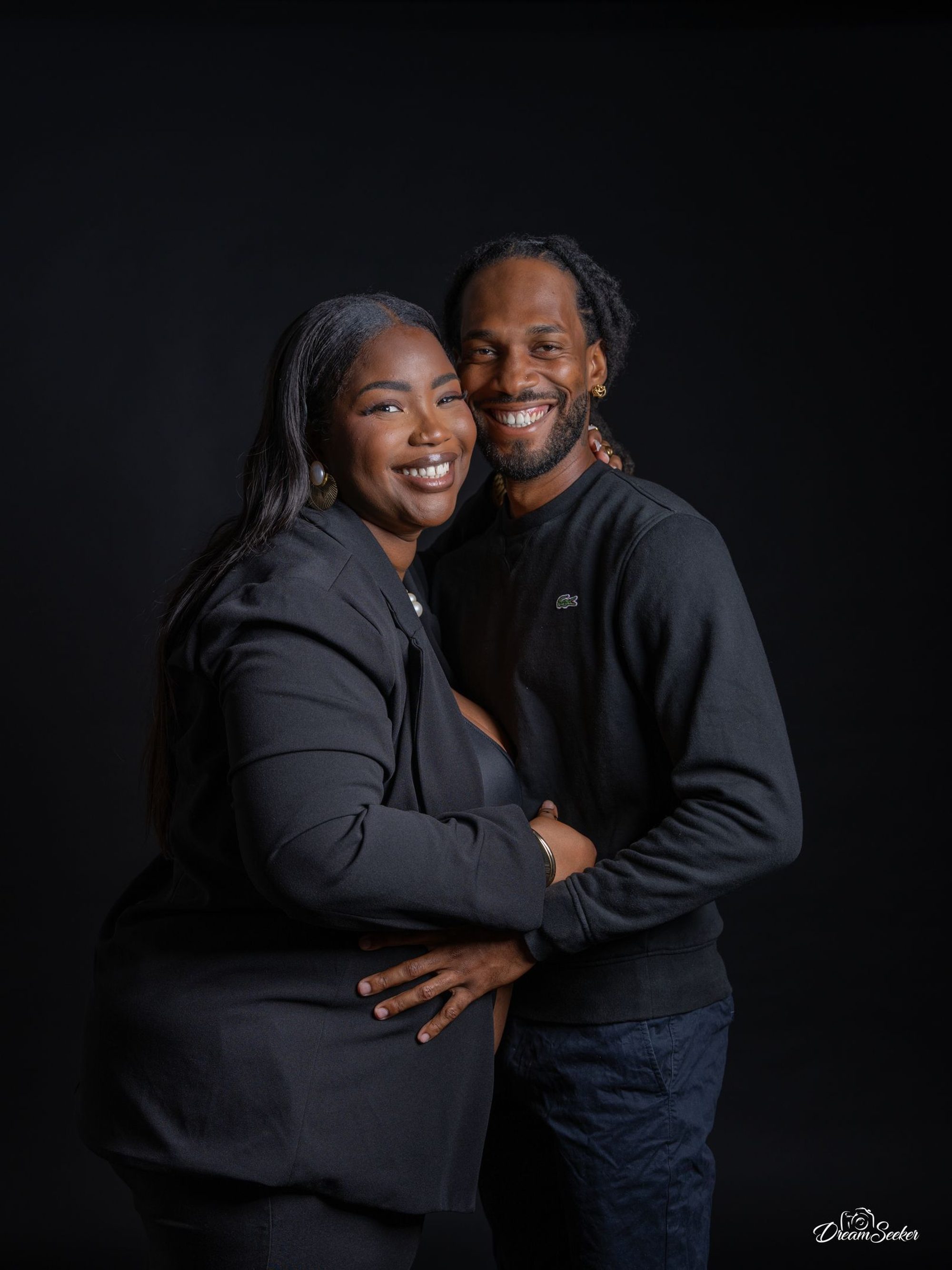 Couple souriant en tenue noire sur fond noir, main sur le ventre