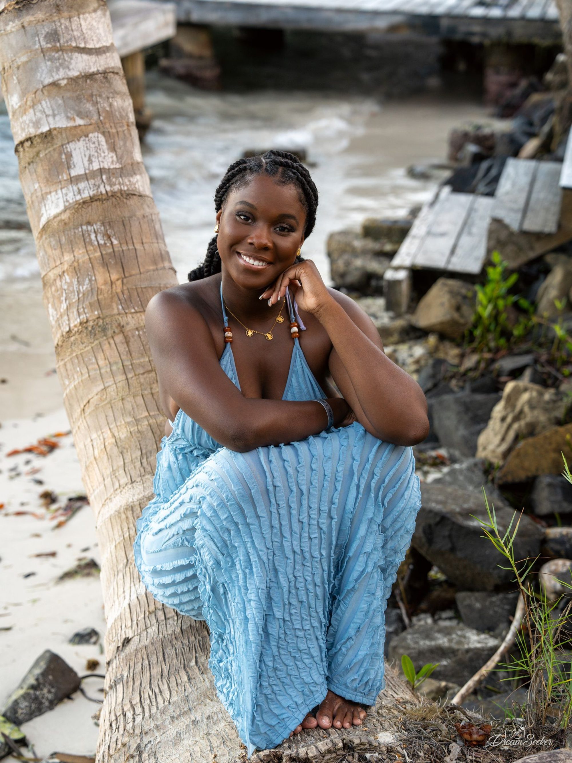 Femme souriante en robe bleue assise contre un tronc de cocotier