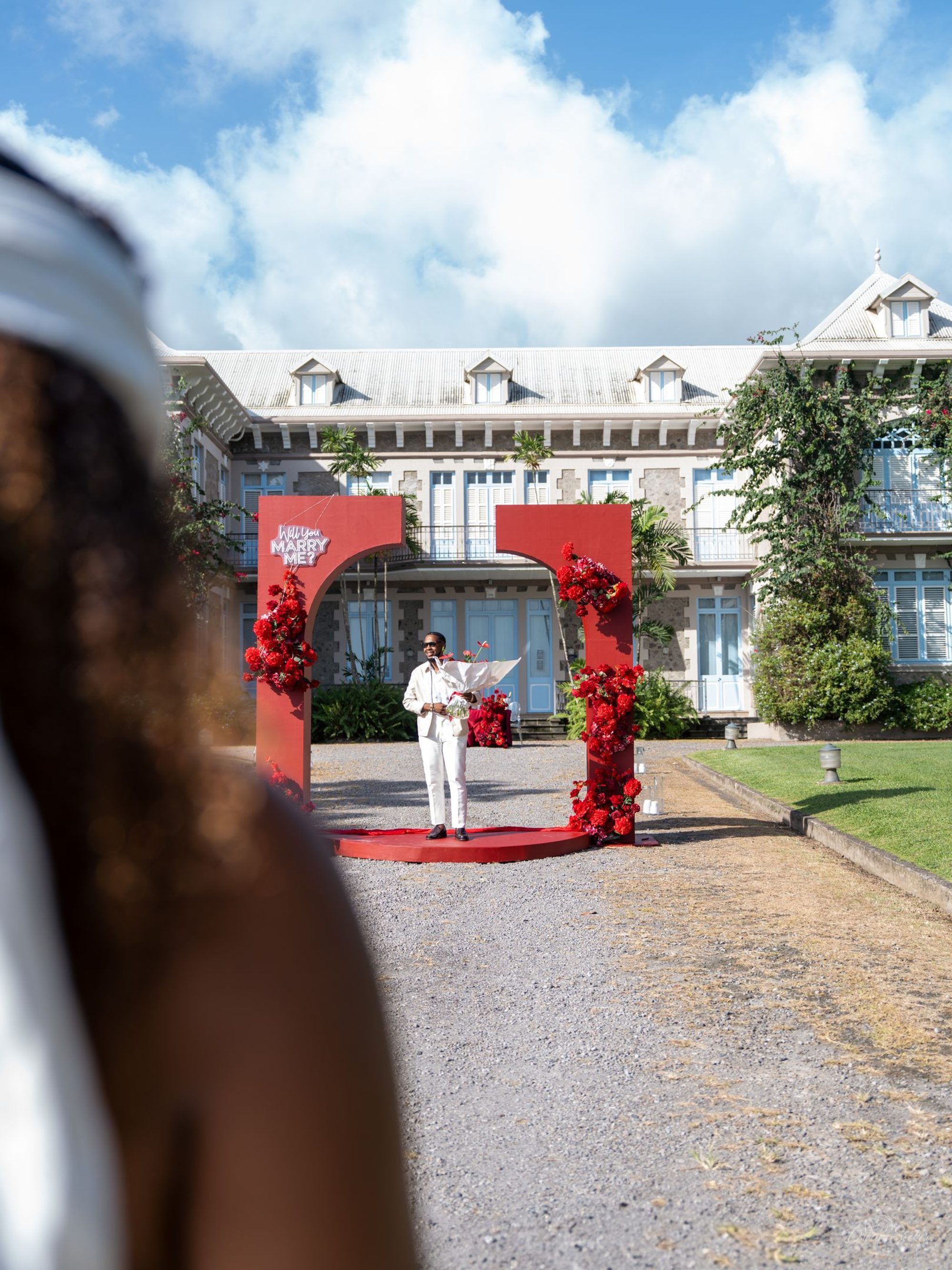 Demande en mariage sous une arche de fleurs rouges, devant une maison créole