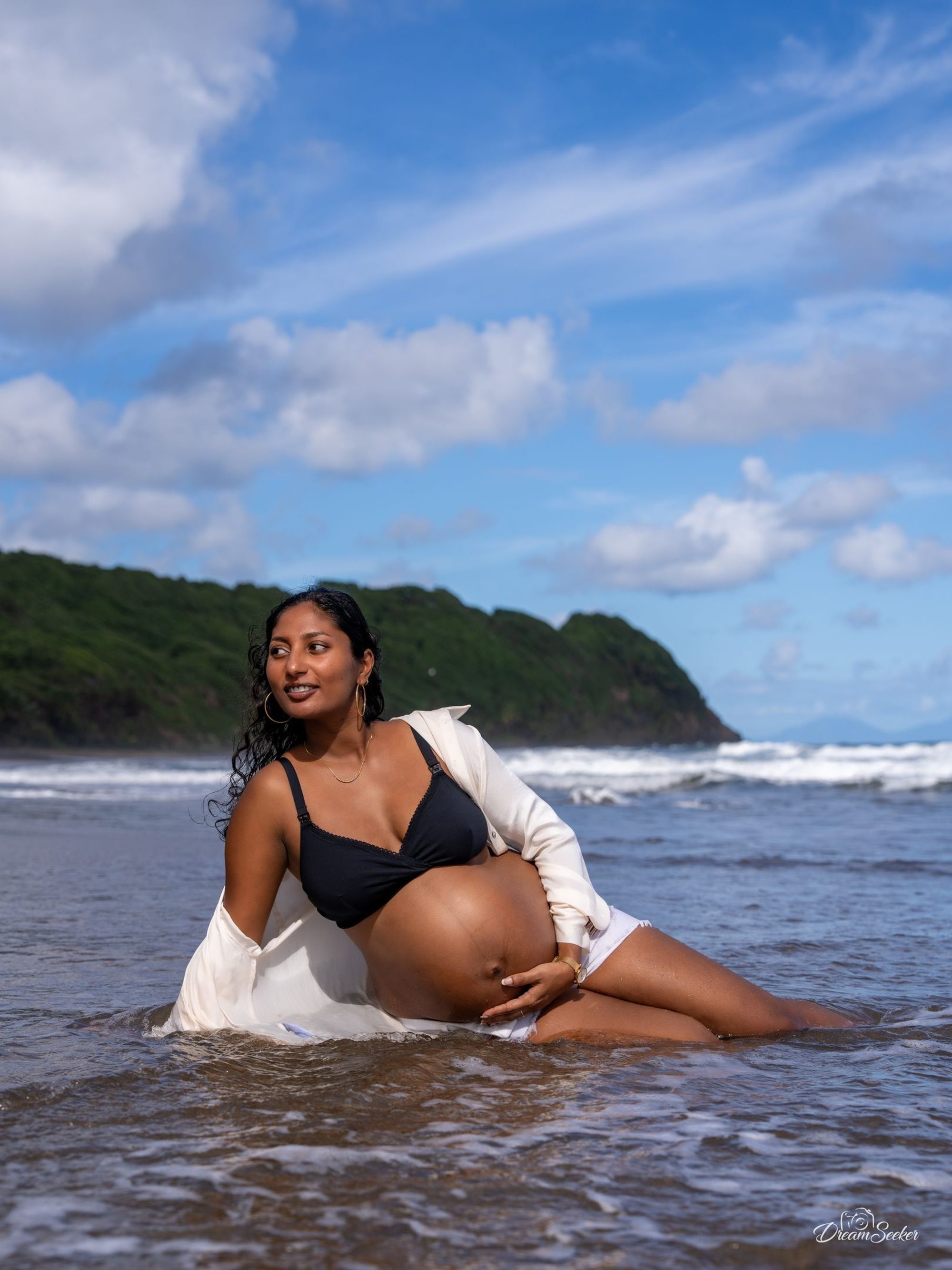 Future mère assise dans l’eau, chemise blanche ouverte, falaise verte