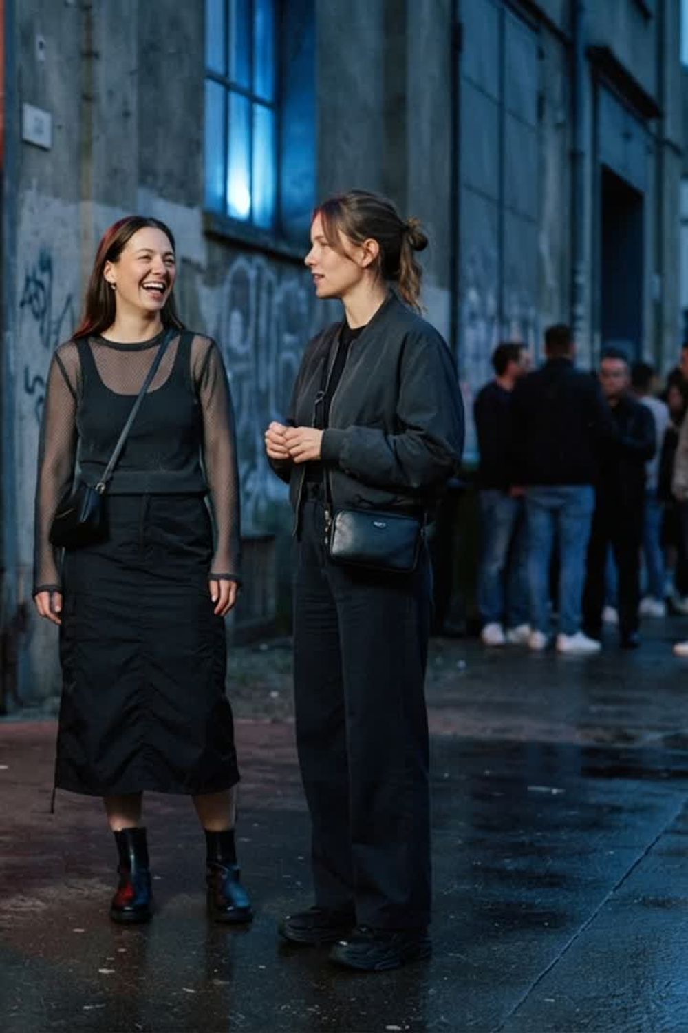 three fictional adults in practical dark outfits outside an anonymous industrial nightlife entrance