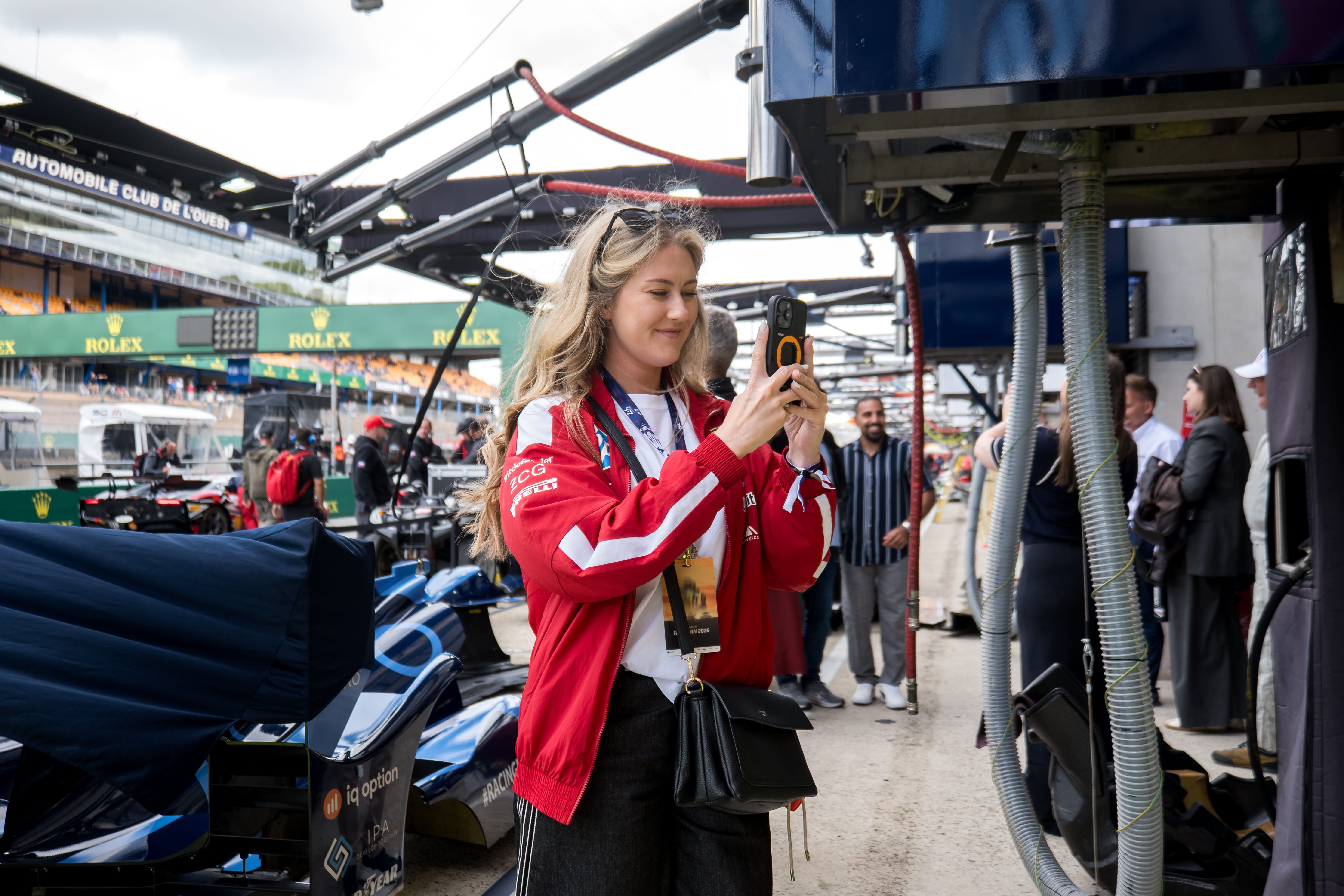 Jeune femme en veste rouge photographiant une voiture de course dans les stands des 24 Heures du Mans